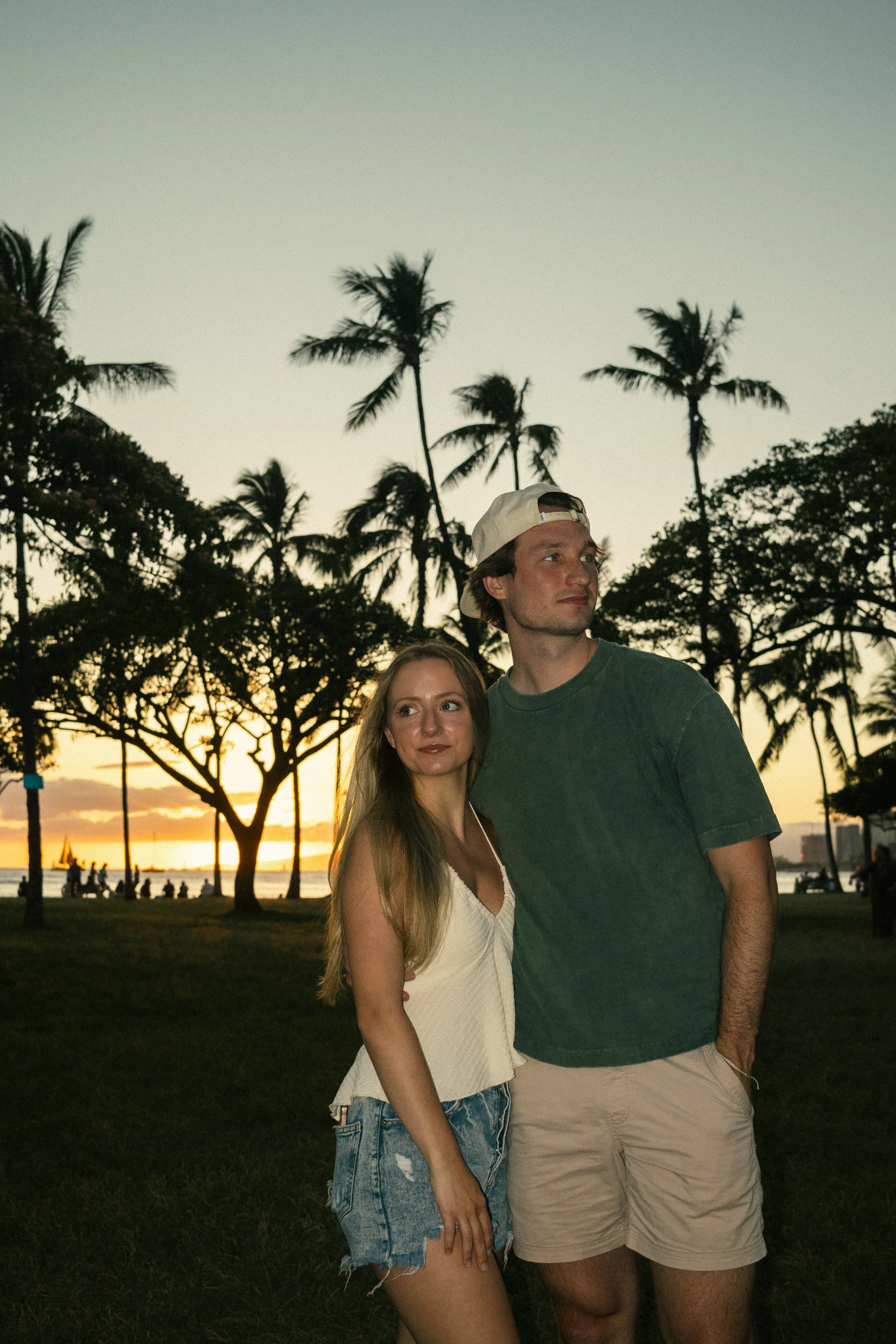 A young couple stands together in a park during sunset, with palm trees and a body of water in the background. The woman has long blonde hair and wears a white sleeveless top and denim shorts. The man wears a green t-shirt, beige shorts, and a white 