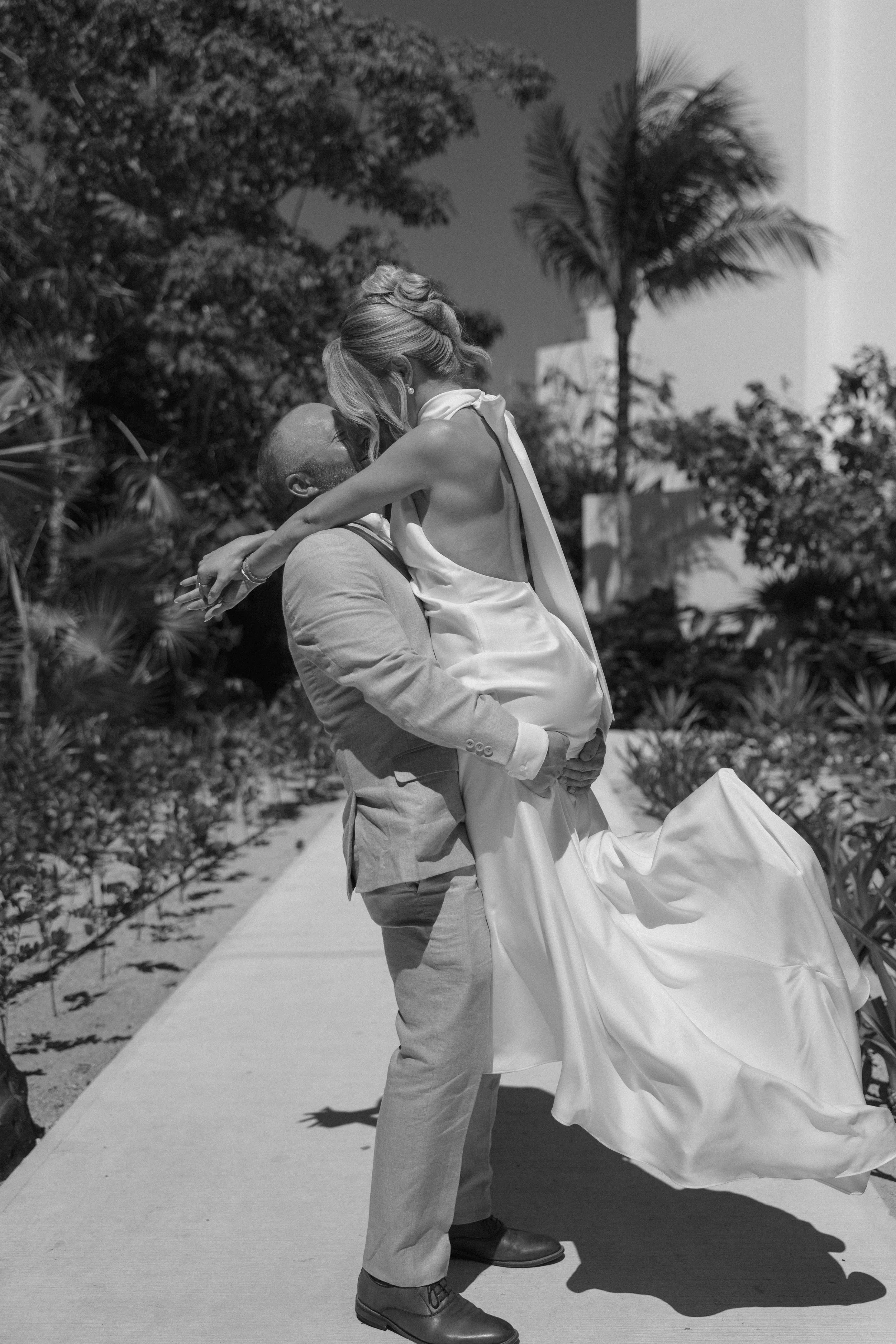 A man in a suit lifting a woman in a wedding dress outdoors, with palm trees and lush plants in the background.