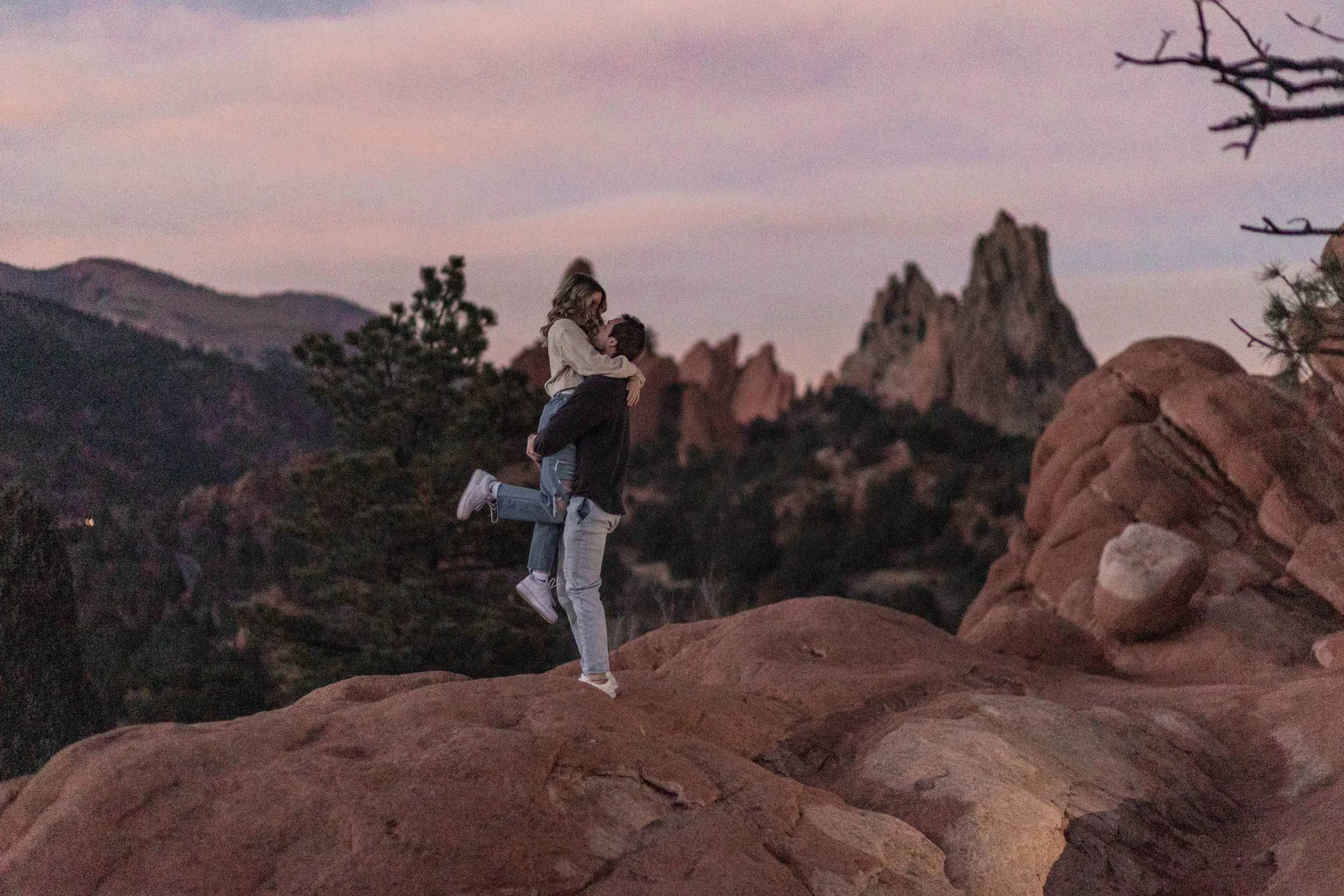 A couple is outdoors at sunset, with one person giving the other a piggyback ride, both smiling. The background features rugged mountains and rocks, with a few leafless branches visible in the foreground.
