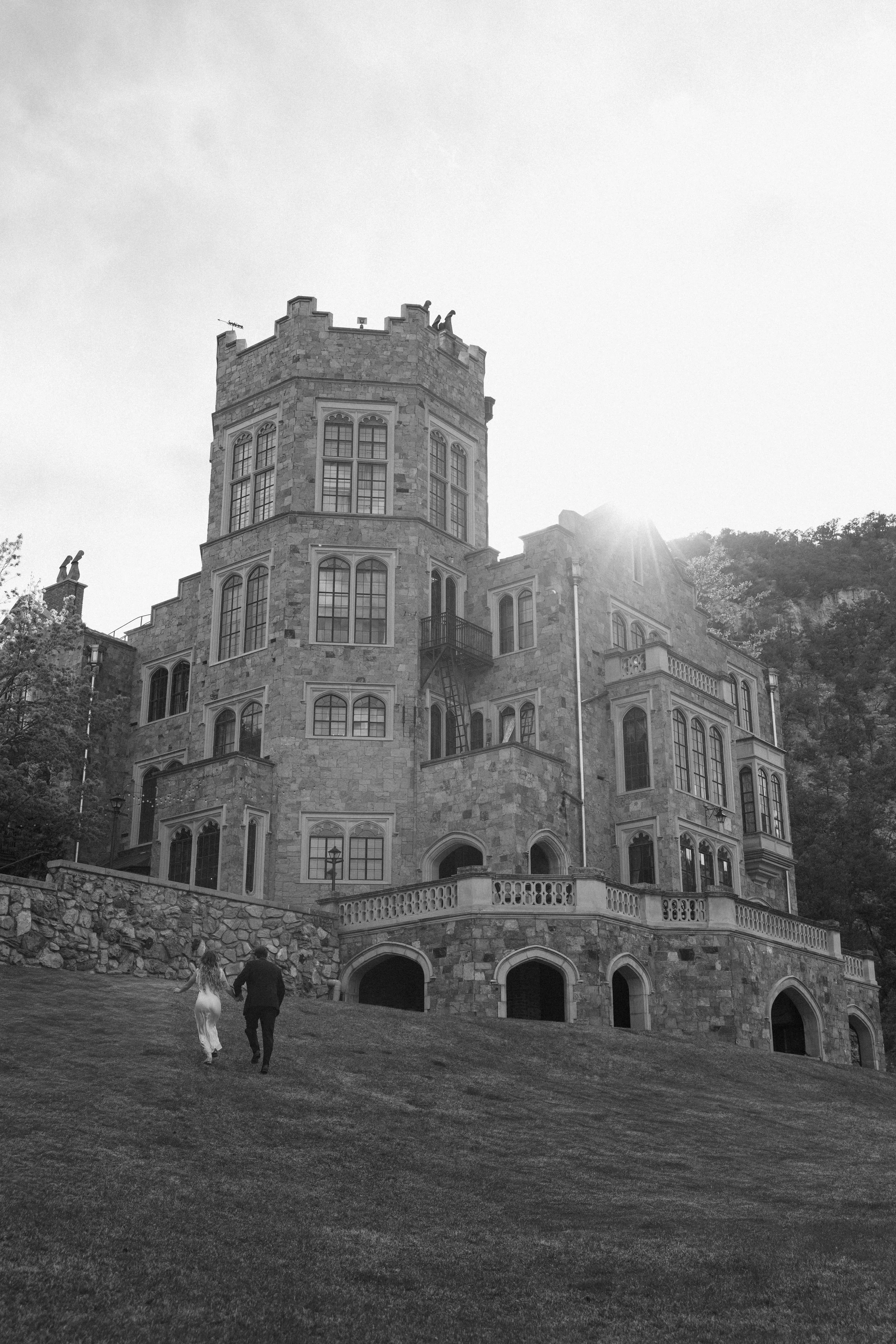 A man and a woman in formal attire walking towards a grand castle-like building made of stone with large windows, balconies, and a tower, with sunlight shining from behind.