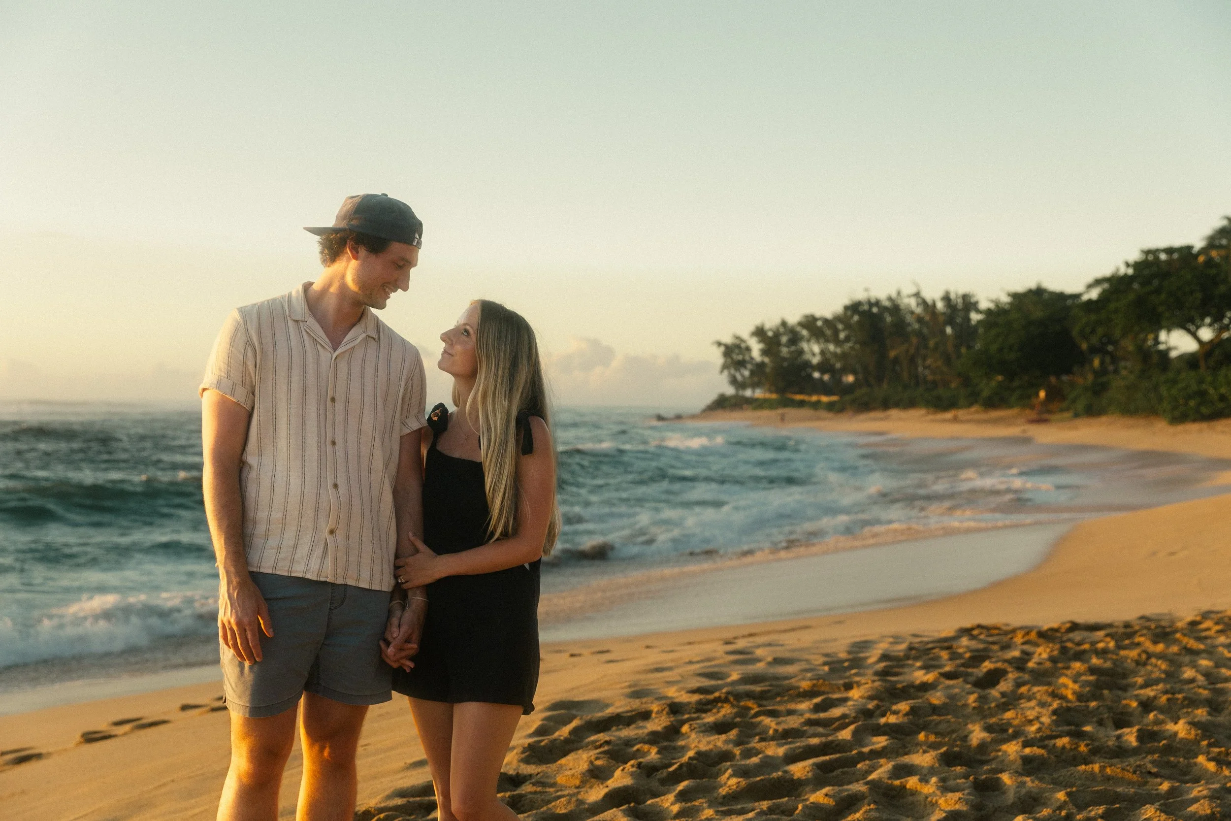 A young couple stands close together on a beach at sunset, holding hands and smiling at each other, with waves and trees in the background.