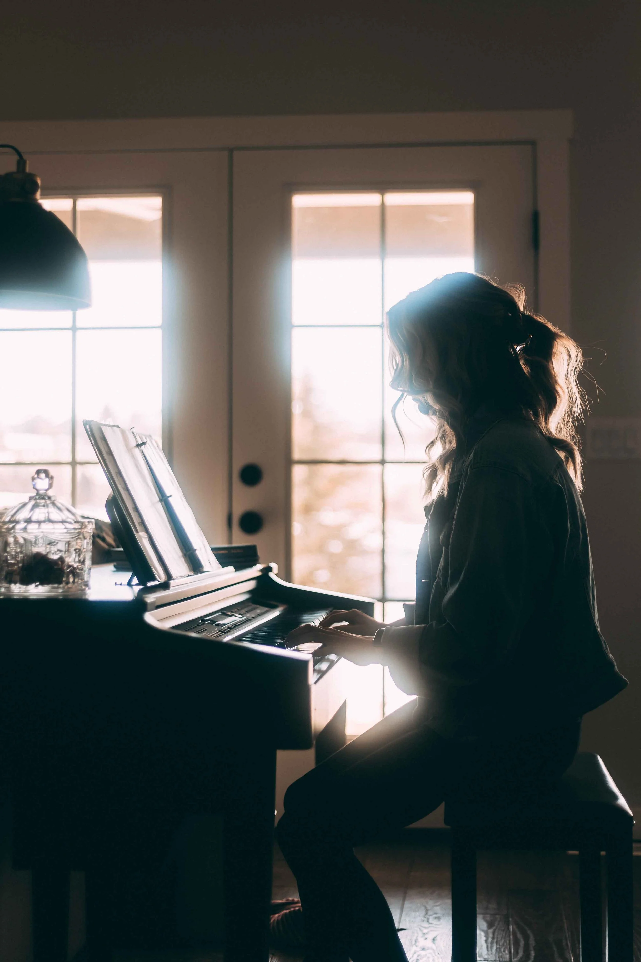 A woman playing the piano in a sunlit room, with sunlight streaming through large windows behind her, creating a warm and peaceful atmosphere.