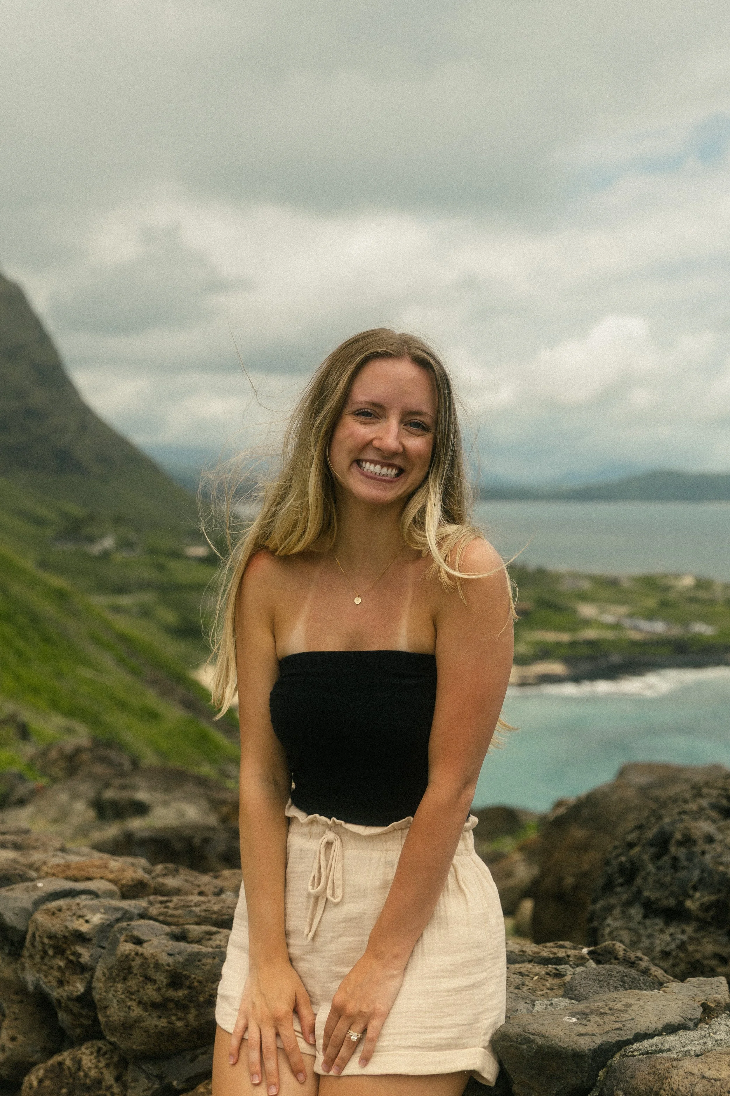 A young woman with long blonde hair and a bright smile standing outdoors on rocks near a coastline with green hills and mountains in the background.