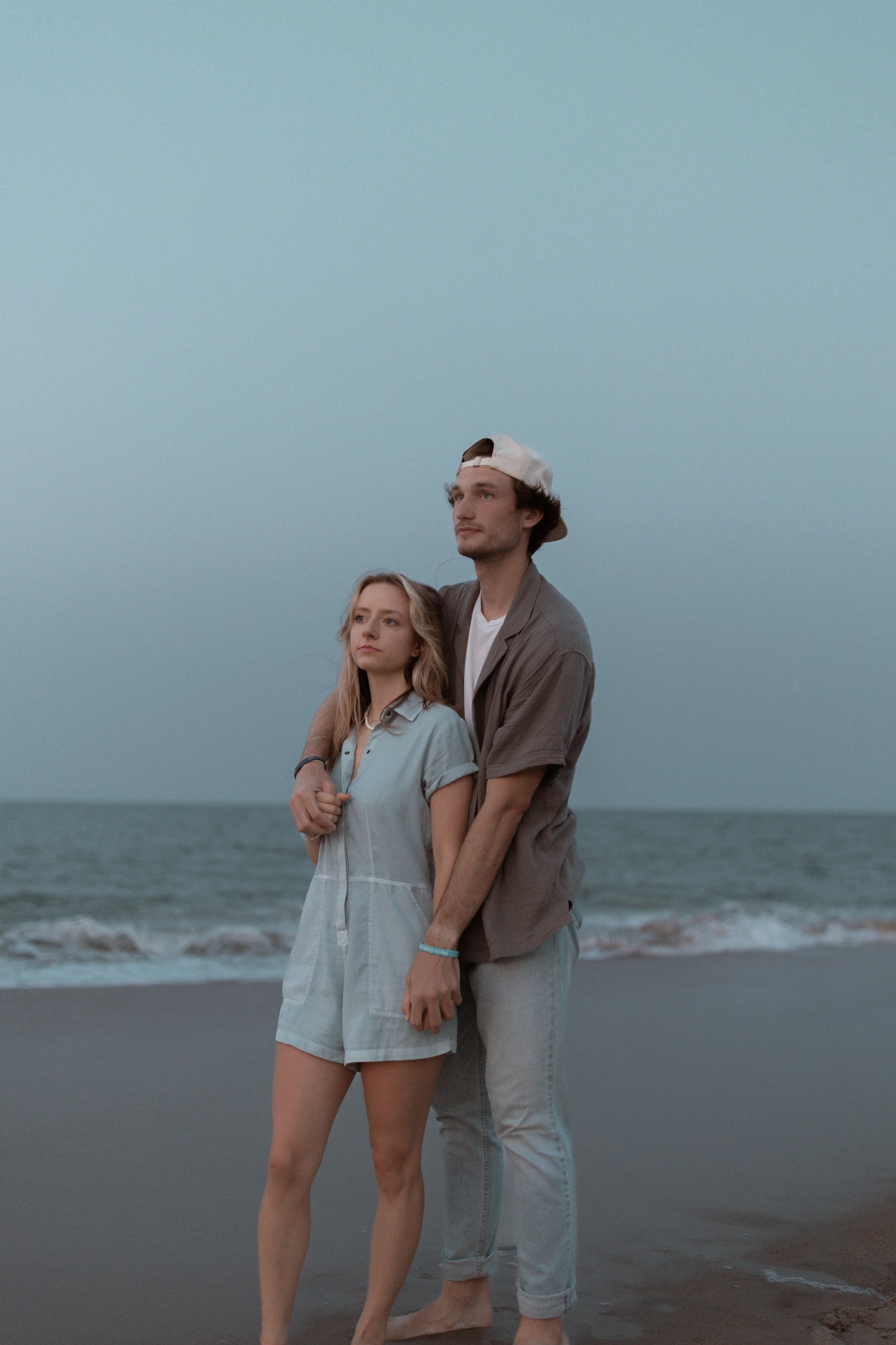 A young man and woman stand close together on a beach at sunset, with the ocean in the background. The man is wearing a backwards white baseball cap, a gray shirt, and light-colored jeans, while the woman is wearing a light blue dress. They are holdi