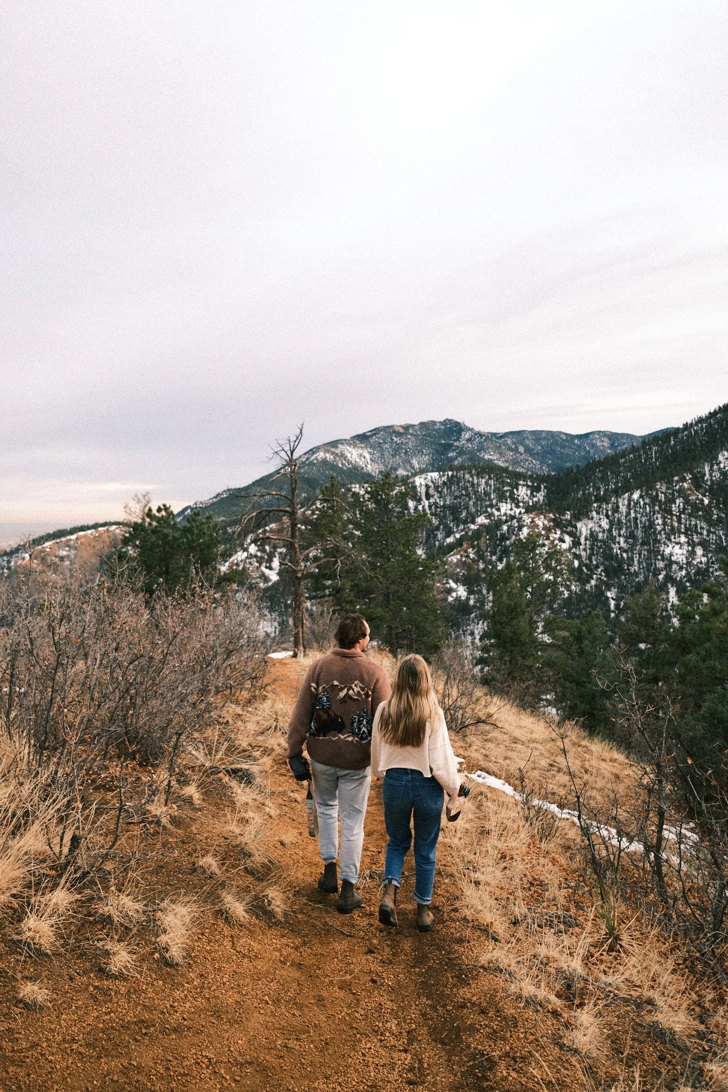 A man and a woman walking on a dirt trail through a mountainous landscape with sparse trees, some snow on the ground, and a cloudy sky.