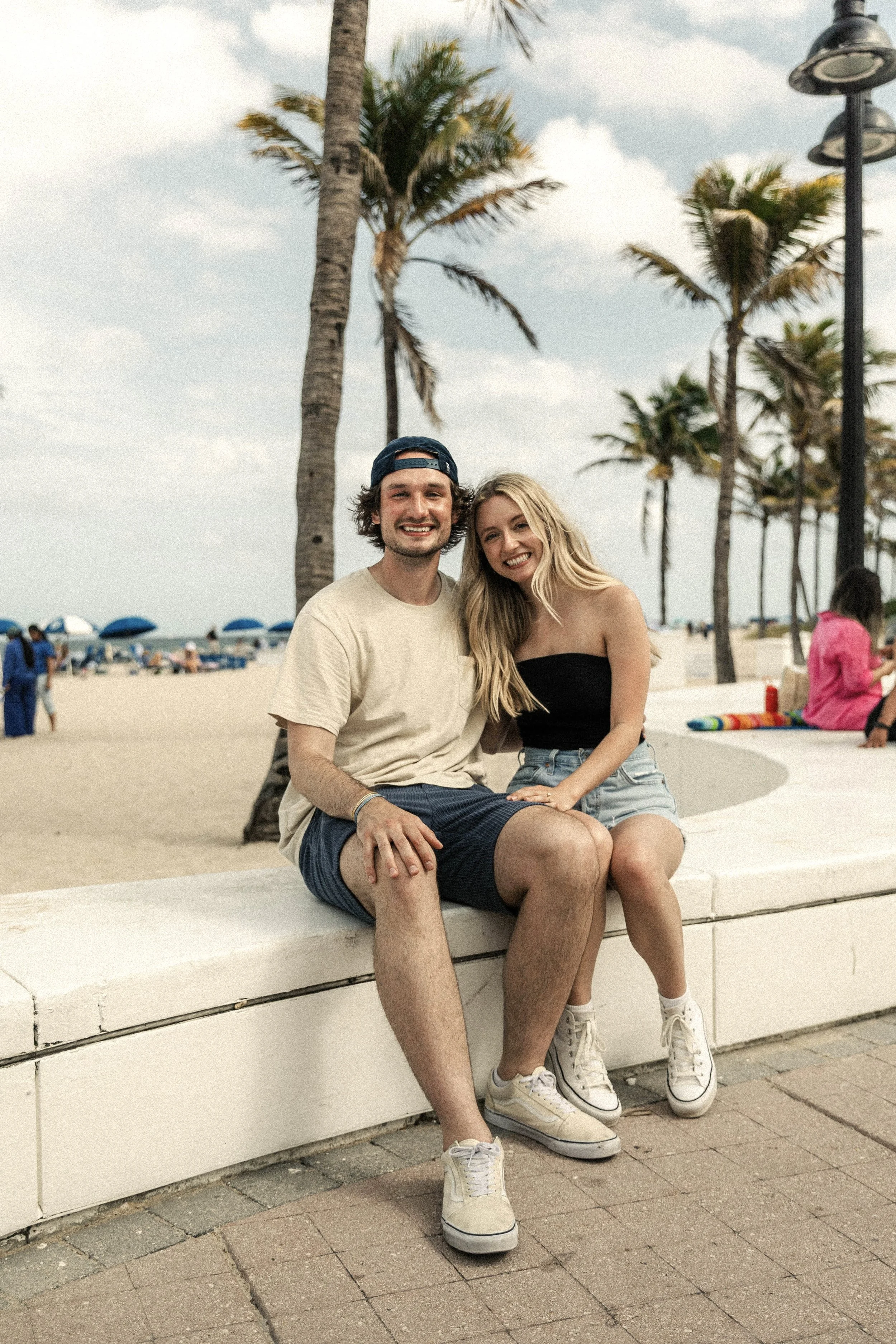 A young man and woman sit close together on a white bench at the beach, smiling at the camera. The man is wearing a beige T-shirt, dark shorts, and a blue cap worn backwards. The woman has long blonde hair, a black strapless top, and denim shorts. In