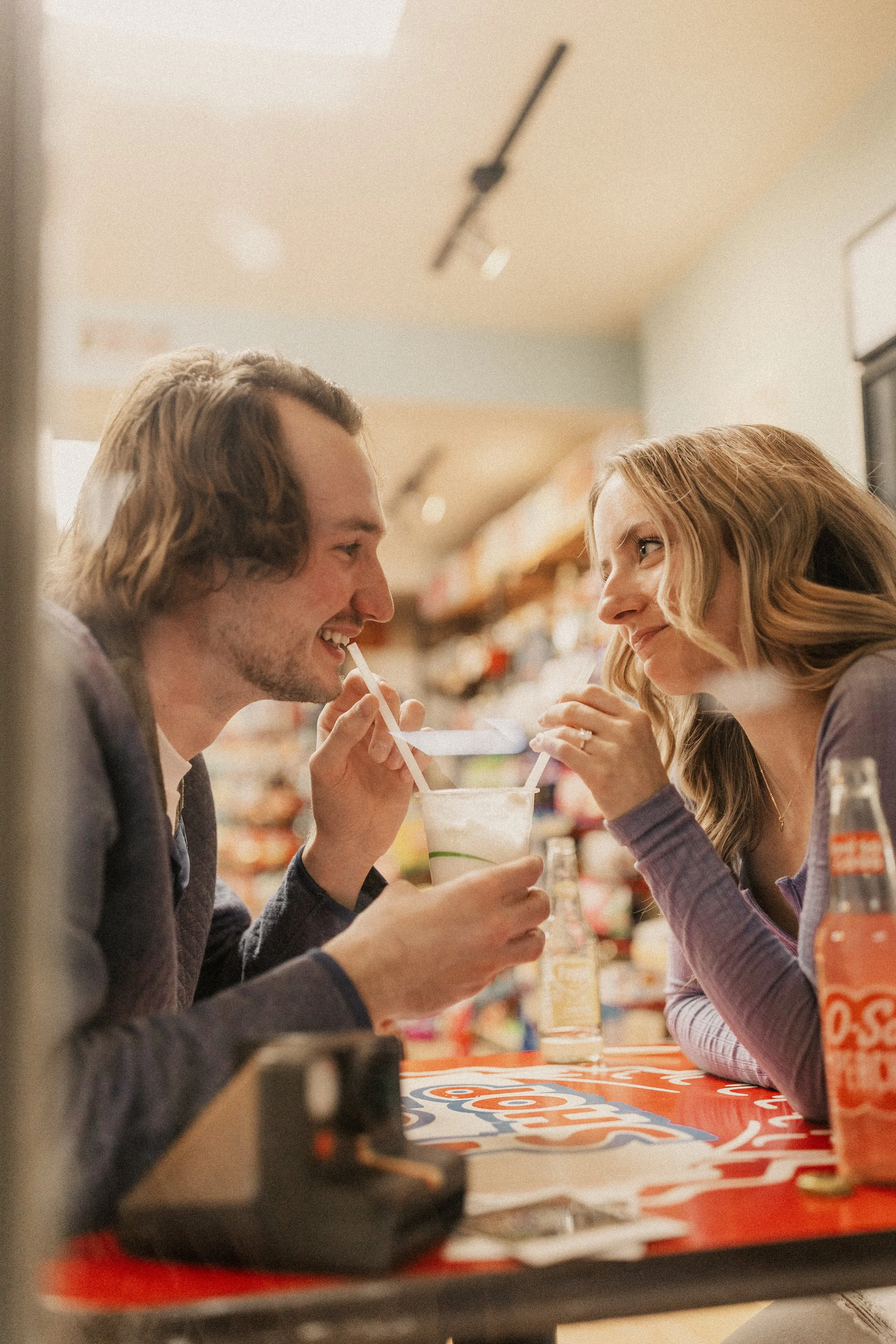 A man and woman at a fast food restaurant counter, sharing a drink with straws, smiling at each other.