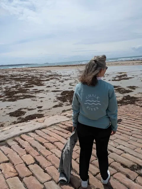 Woman standing on a brick path near the beach, facing towards the ocean, with gray sky overhead.