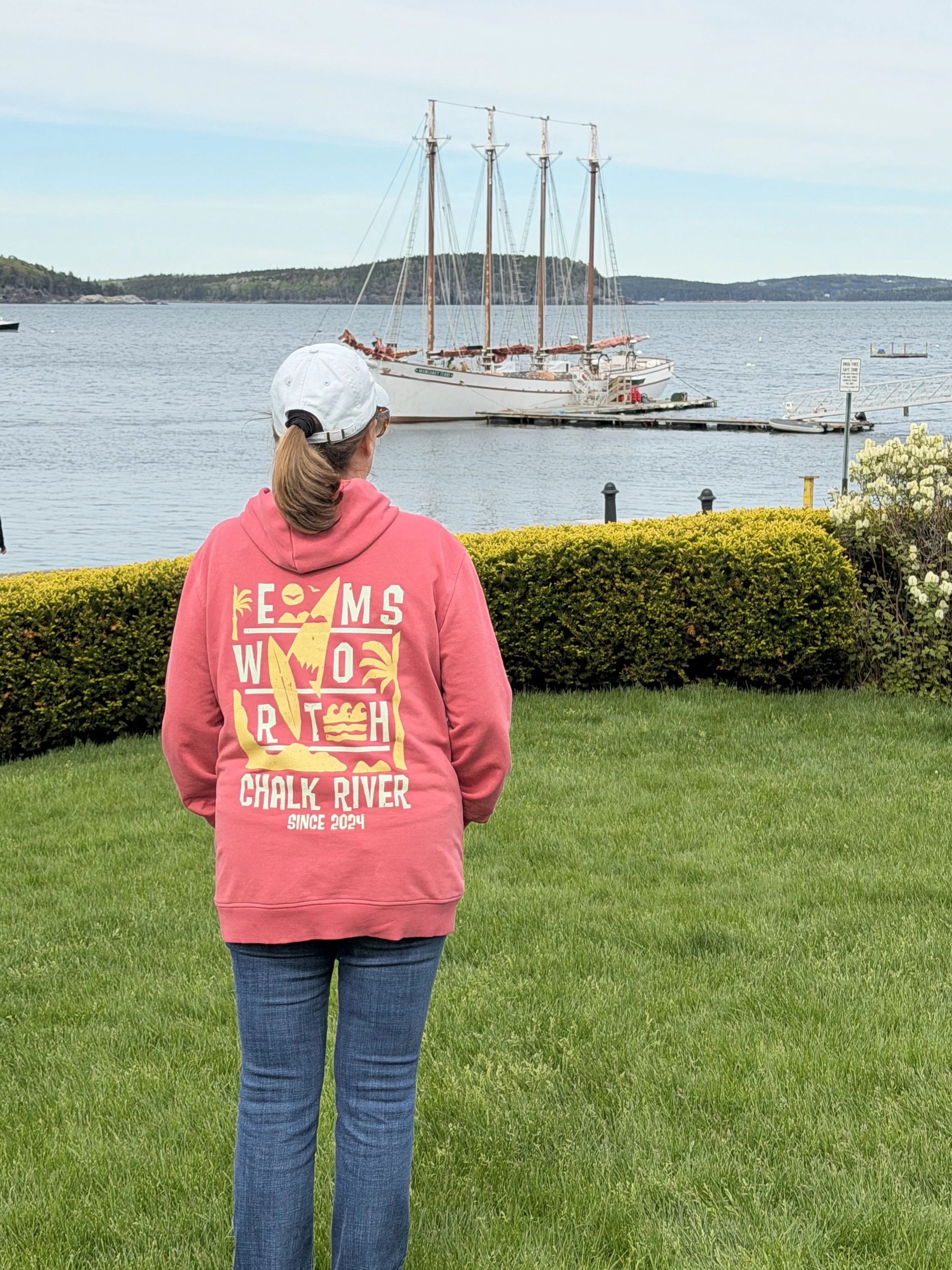 A woman with a ponytail, wearing a white cap and a pink hoodie that reads 'TEAMS WO RCH CHALK RIVER SINCE 2024,' is standing on green grass in front of a lake, observing a wooden sailing ship docked at a pier with distant hills and a cloudy sky in th