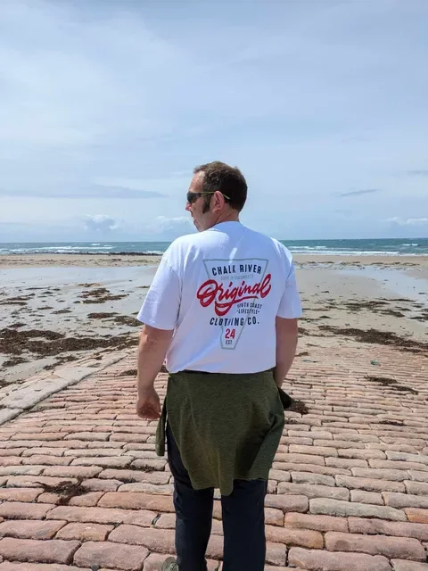 A man stands on a brick pathway facing the beach, wearing sunglasses, a white graphic t-shirt, and dark pants, with a jacket tied around his waist.
