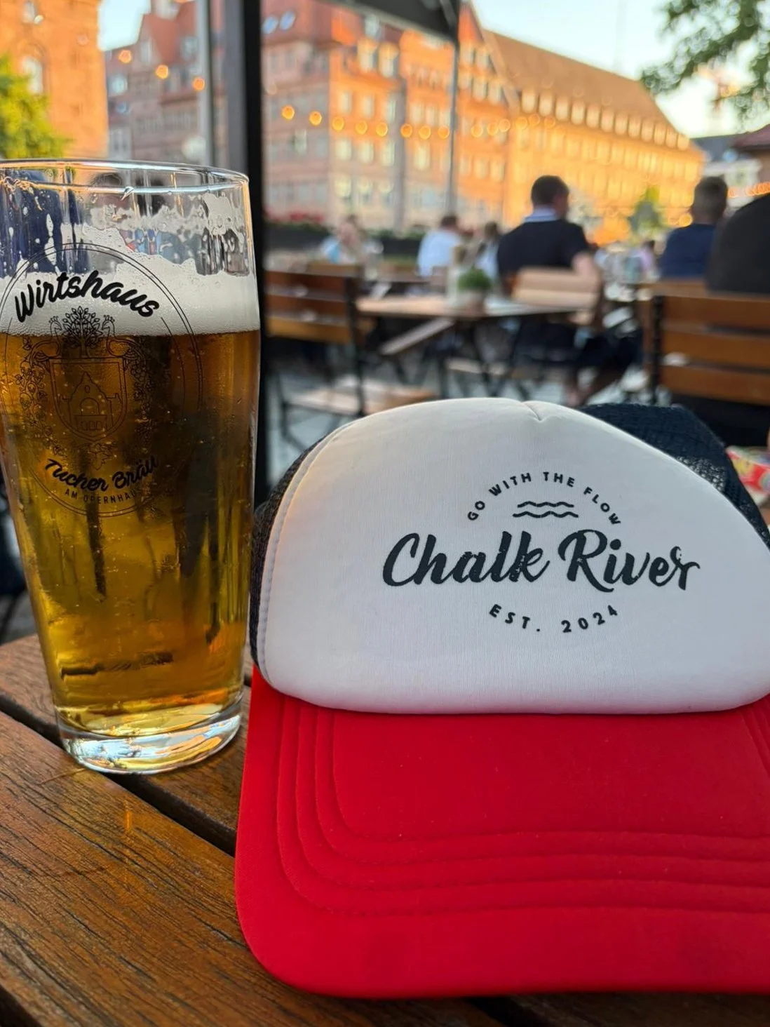 A glass of beer and a white and red baseball cap on a wooden table in an outdoor restaurant or bar with people sitting at tables. The background shows a cityscape with historic buildings and string lights.