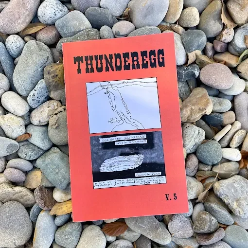 A red booklet titled "THUNDEREGG" with black bold letters, placed on a bed of multicolored smooth stones. The cover features two black-and-white diagrams of geological formations.