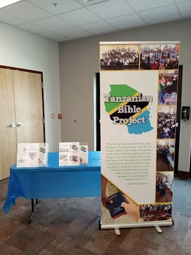A vertical banner for the Tanzanian Bible Project with images of groups and activities, positioned next to a table with informational pamphlets on a blue tablecloth.