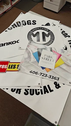 A pile of printed signs and posters on a table, including a 'Dogs' sign, a circular logo with 'Montana' and '406', and a 'Park Street Printers' flyer with colored triangles.