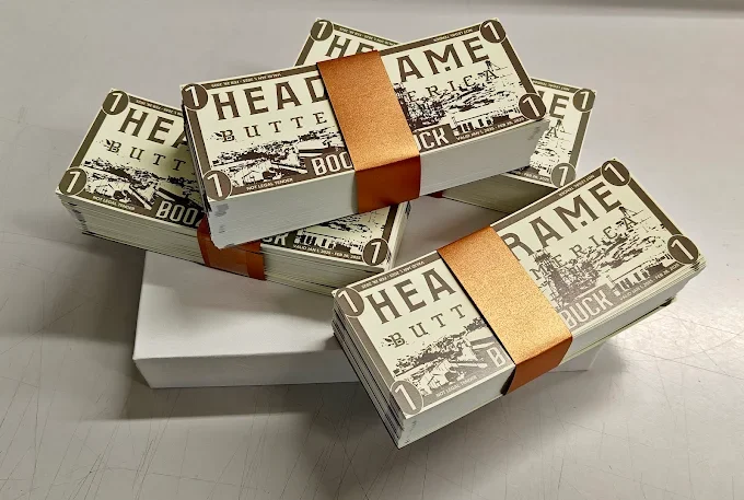 Stacks of paper bills shaped like railway tickets with the text 'HEADRAME BUTT' and the image of a train, wrapped with orange bands, placed on a white surface.