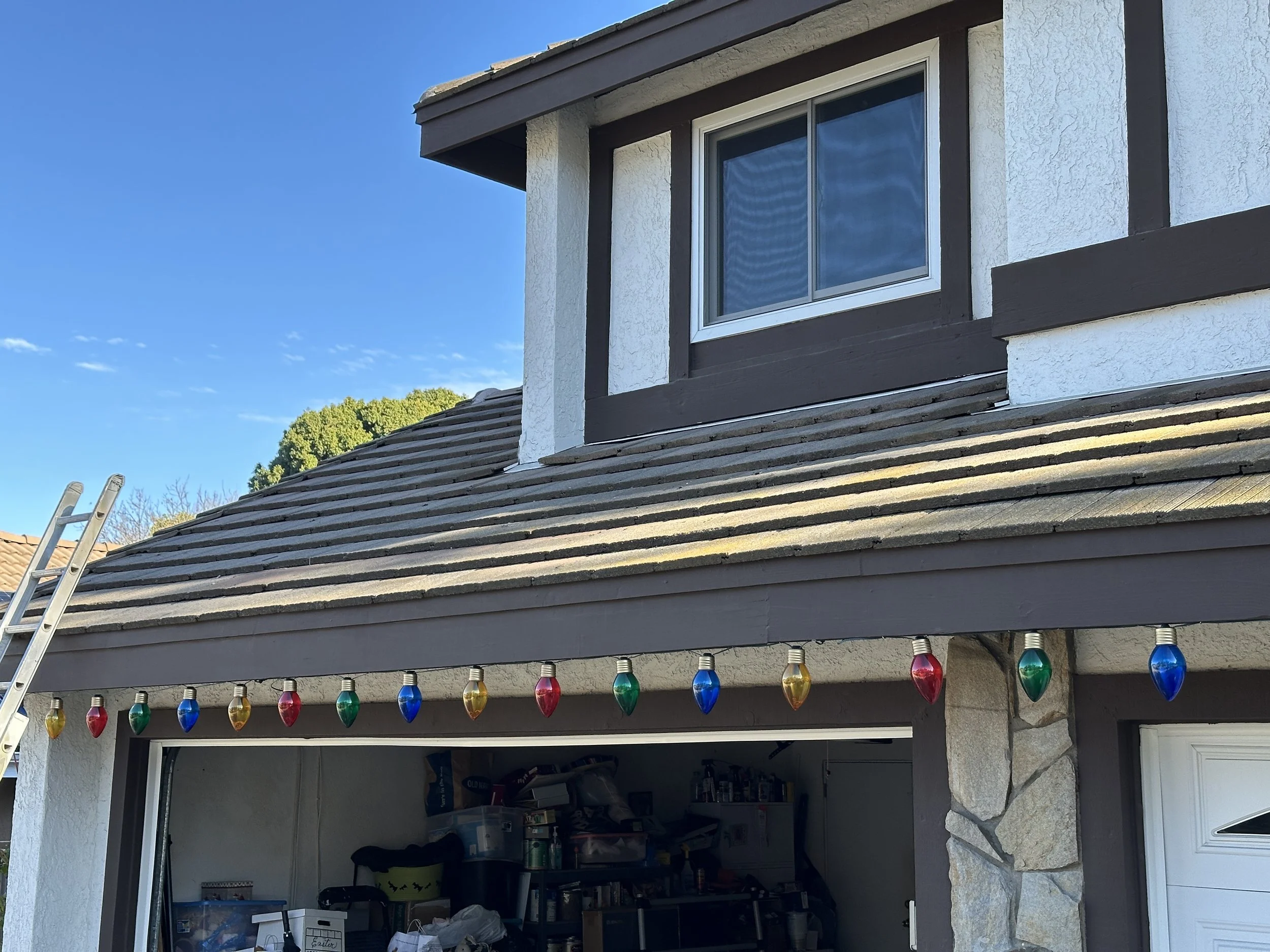 String of colored holiday lights hanging above the garage door of a house with a clamshell roof and a small window on the upper floor.