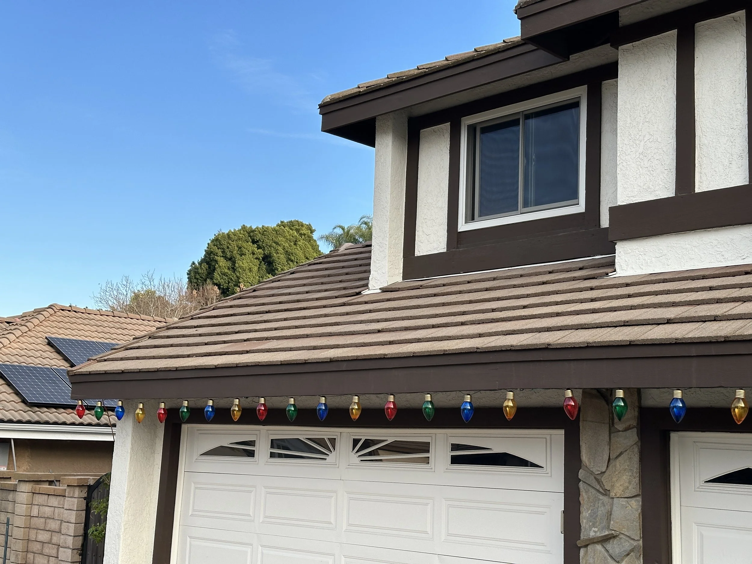 A two-story house with a brown tiled roof, white and brown exterior walls, and a window in the upper floor. Colorful string lights with red, green, blue, and yellow bulbs are hanging along the edge of the roof. Solar panels are visible on the neighbo