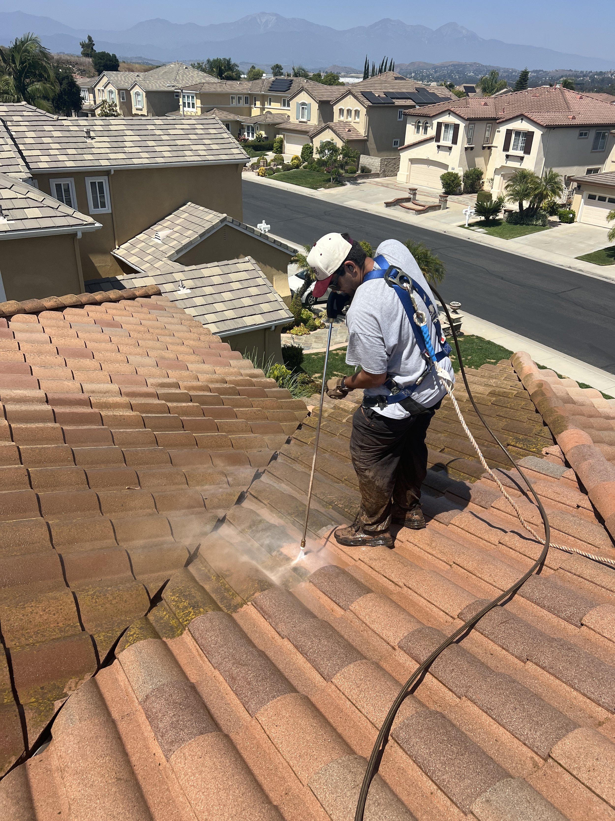 A worker using a pressure washer to clean a tiled roof on a residential house with neighboring houses and mountains in the background.