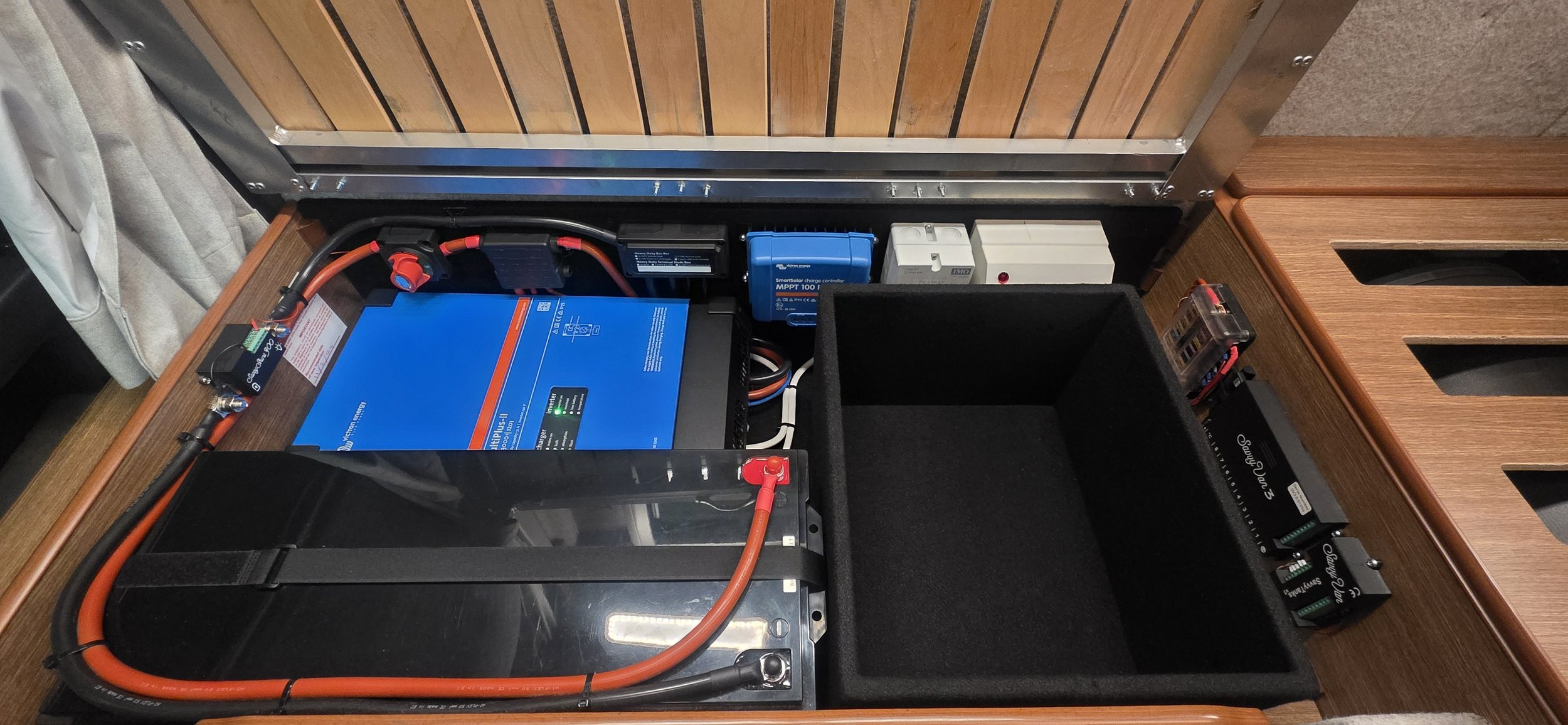 An organized electronics setup featuring a blue inverter, batteries, wiring, and an empty black storage bin, all housed within a wooden case or enclosure.