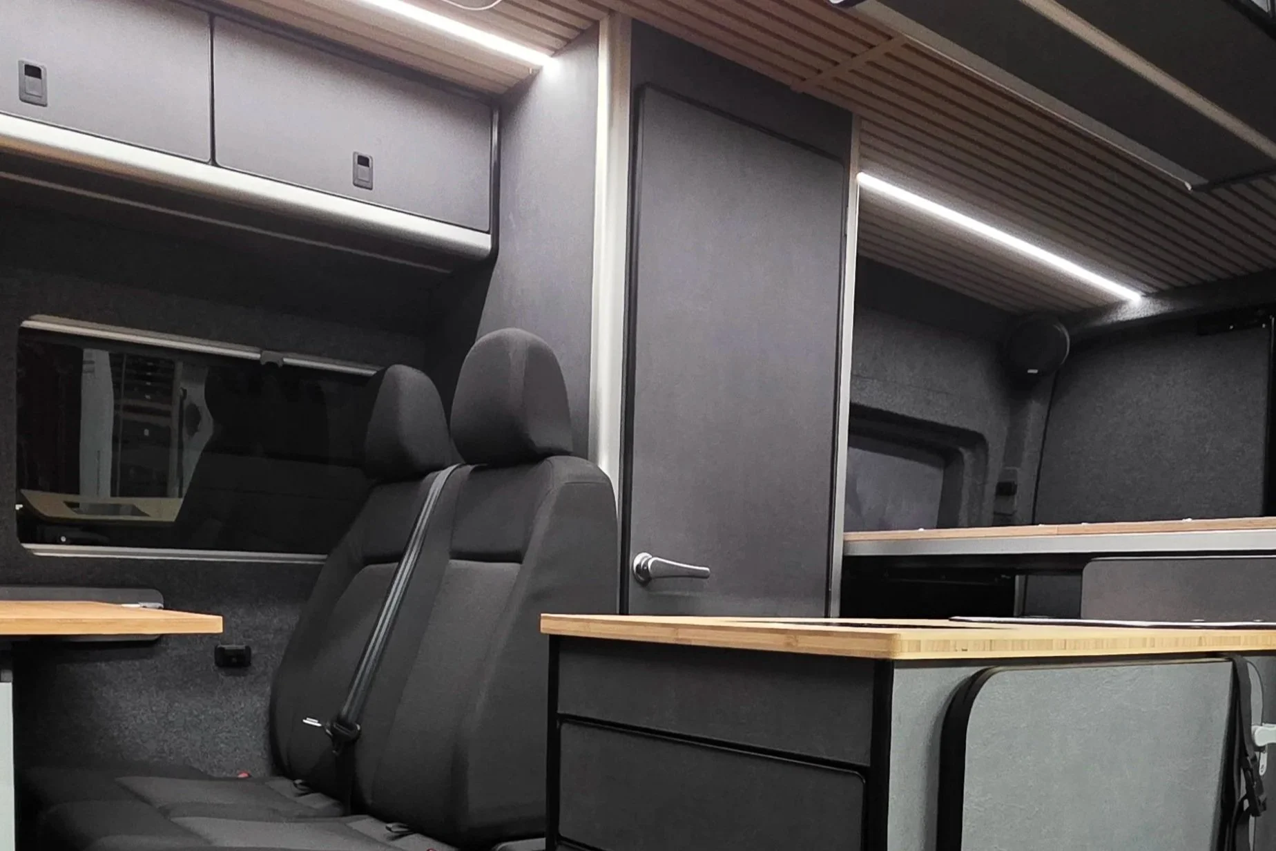Interior of a modern camper or van with black seats, wood countertops, and gray walls and ceiling.