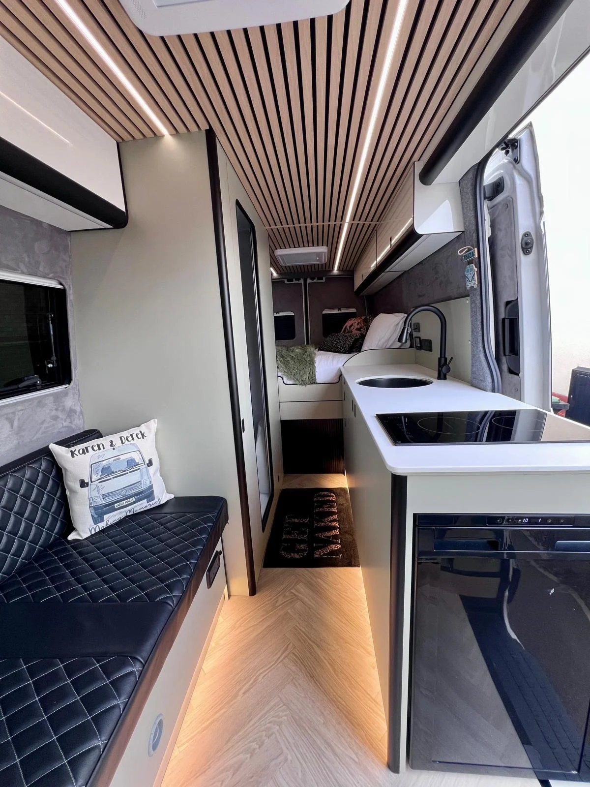 Interior of a modern camper van with a small kitchenette including a sink and stove, a seating area with a quilted black bench and decorative pillows, and a sleeping area with a bed and pillows, all featuring wood accents and sleek design.