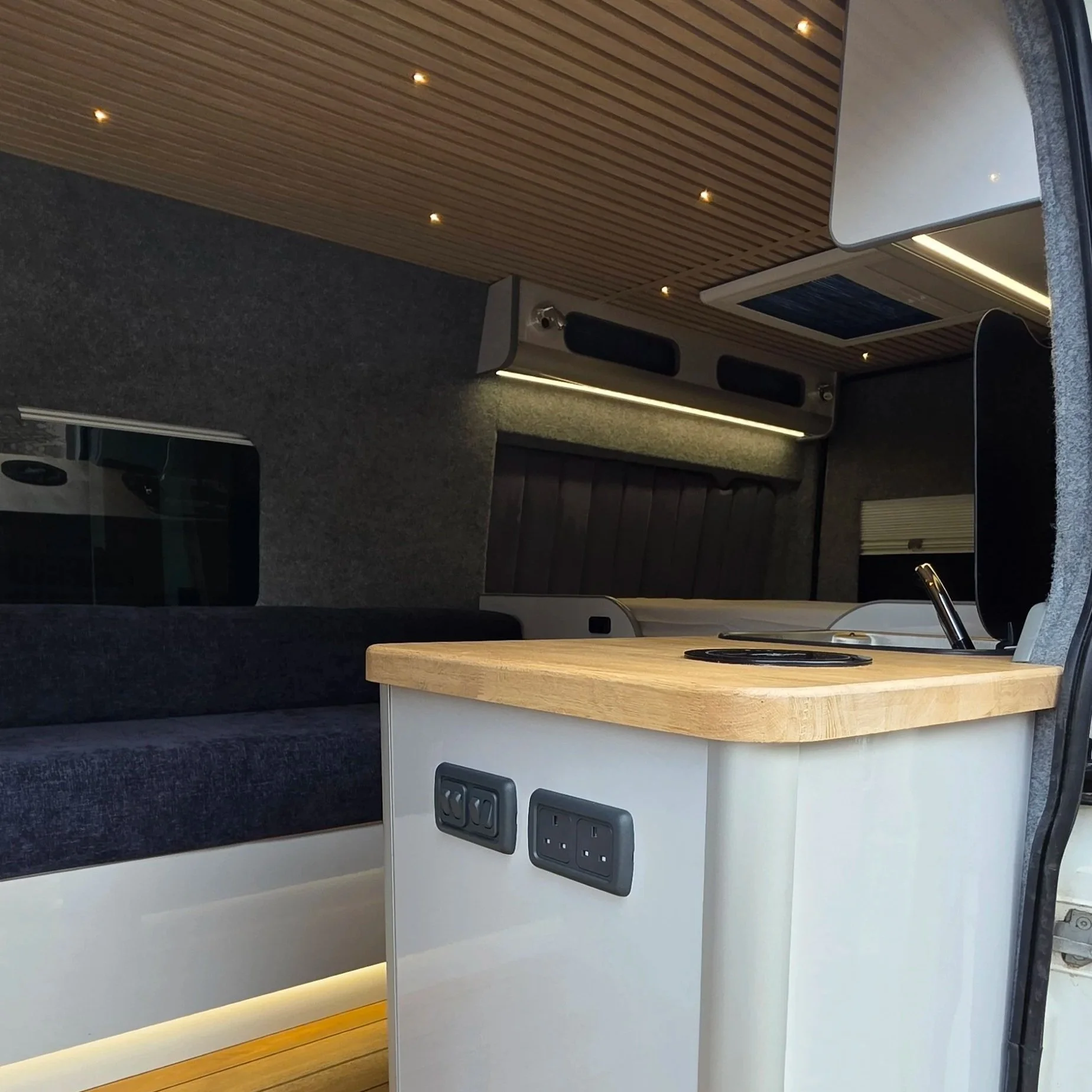Interior of a modern camper van with a wooden ceiling, dark cushioned seating, a small table with power outlets, and various interior features.