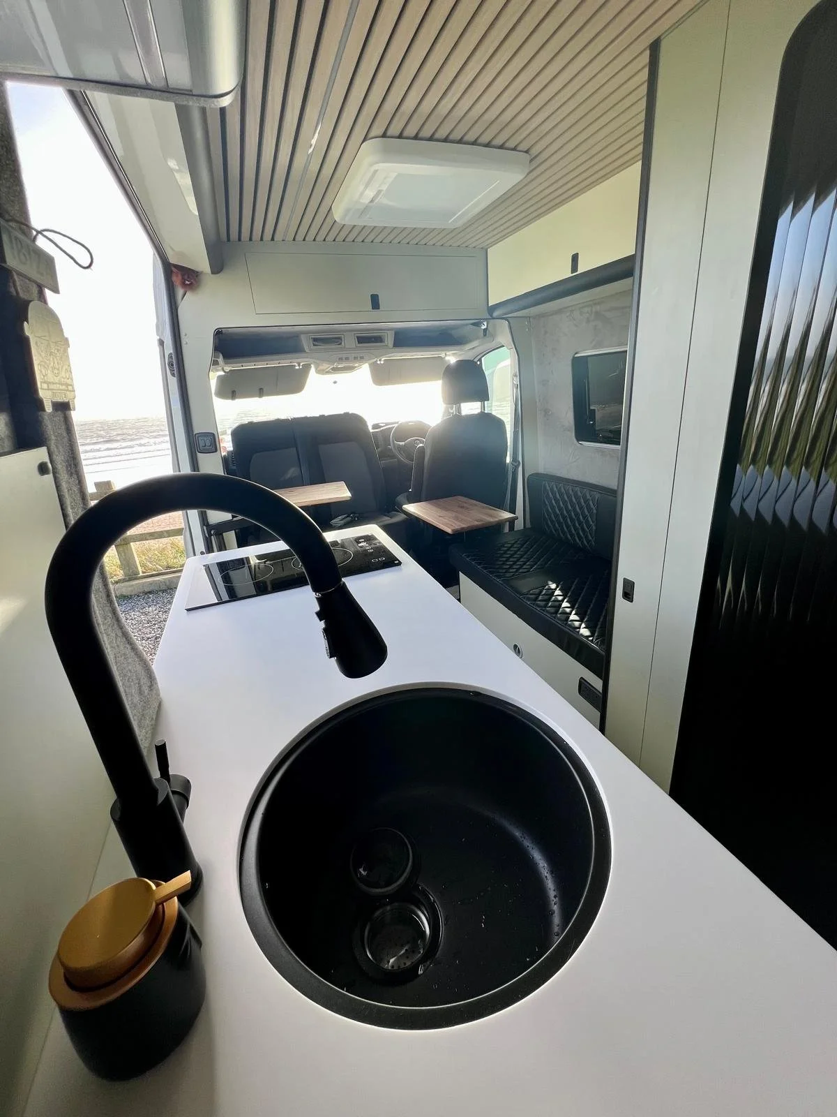 Interior of a camper van with a kitchen area featuring a black sink, a stovetop, and a soap dispenser, with seating and a television visible in the background.