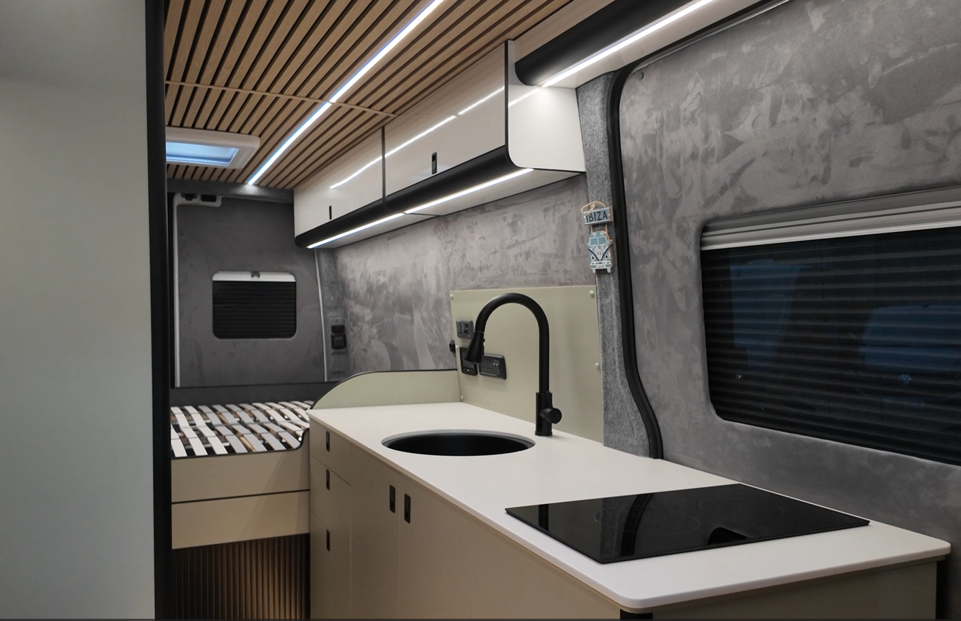 Interior of a modern camper van with a small kitchen area featuring a black sink, a stovetop, and storage cabinets, with a sleeping area in the background and a window with a blind.