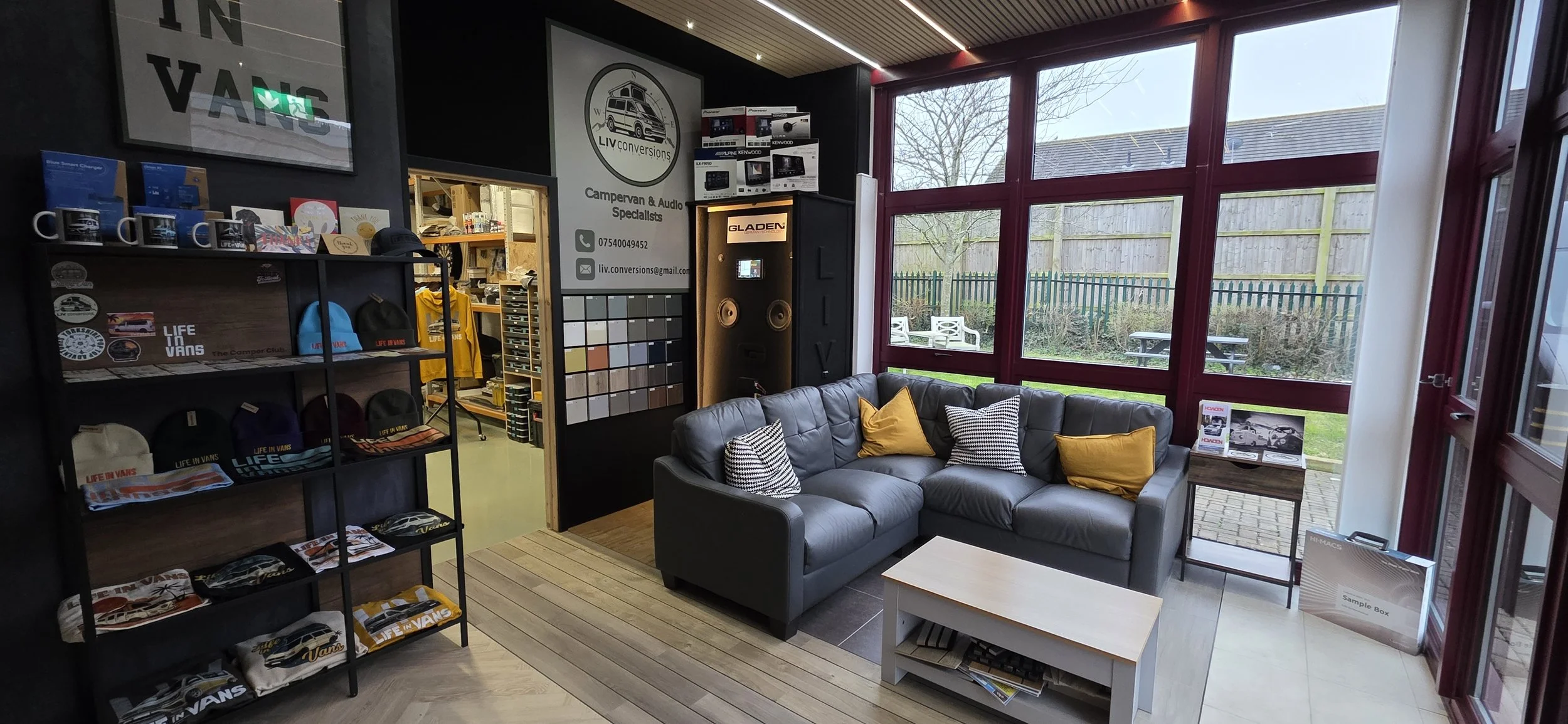 A cozy seating area with a black leather sofa decorated with yellow and black-and-white pillows, situated in a modern showroom with large windows, a small wooden coffee table, and shelves displaying merchandise like mugs, hats, and brochures. Outside, there are benches and tables in a fenced outdoor area.