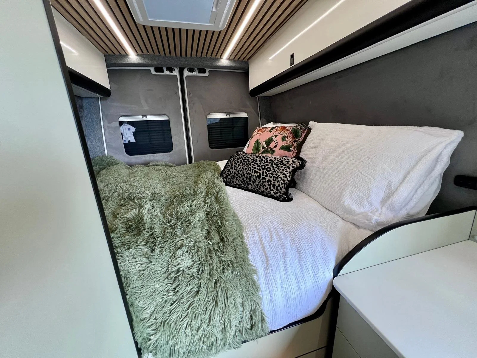 Small sleeping area inside a vehicle or camper, with a bed made up with white sheets, pillows, and a textured green blanket, and a window at the head of the bed.