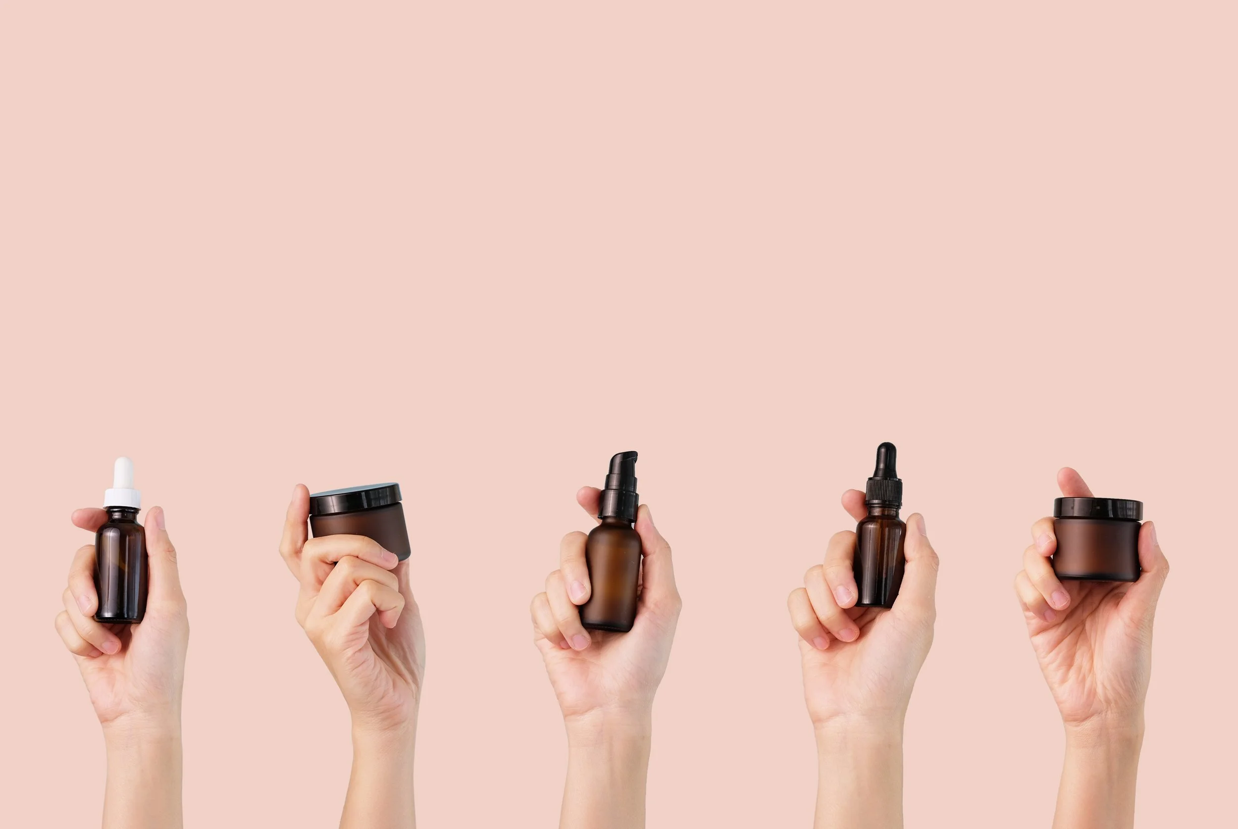 Five hands holding different skincare or cosmetic containers in front of a pink background.