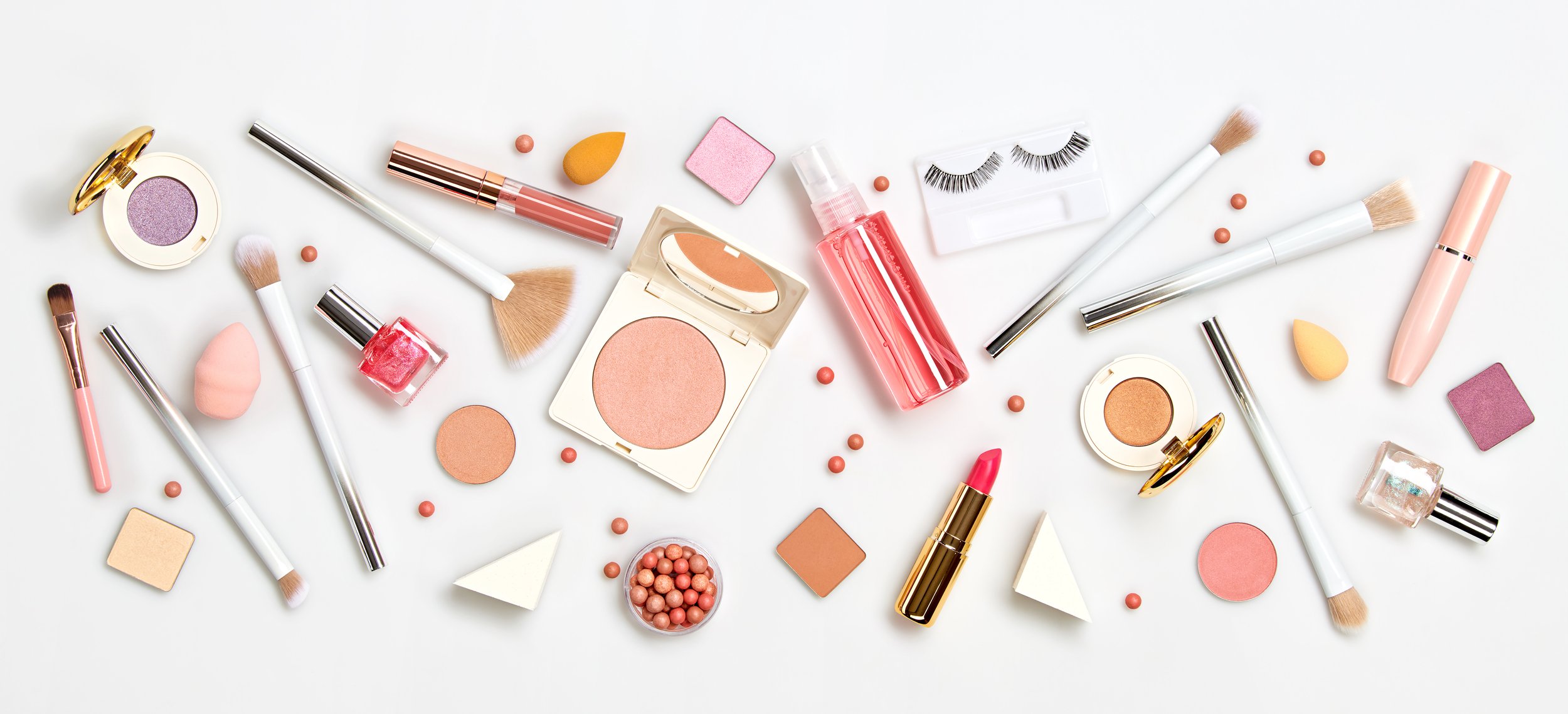 Flat lay of various makeup products including brushes, powders, lipstick, and nail polish on a white background.