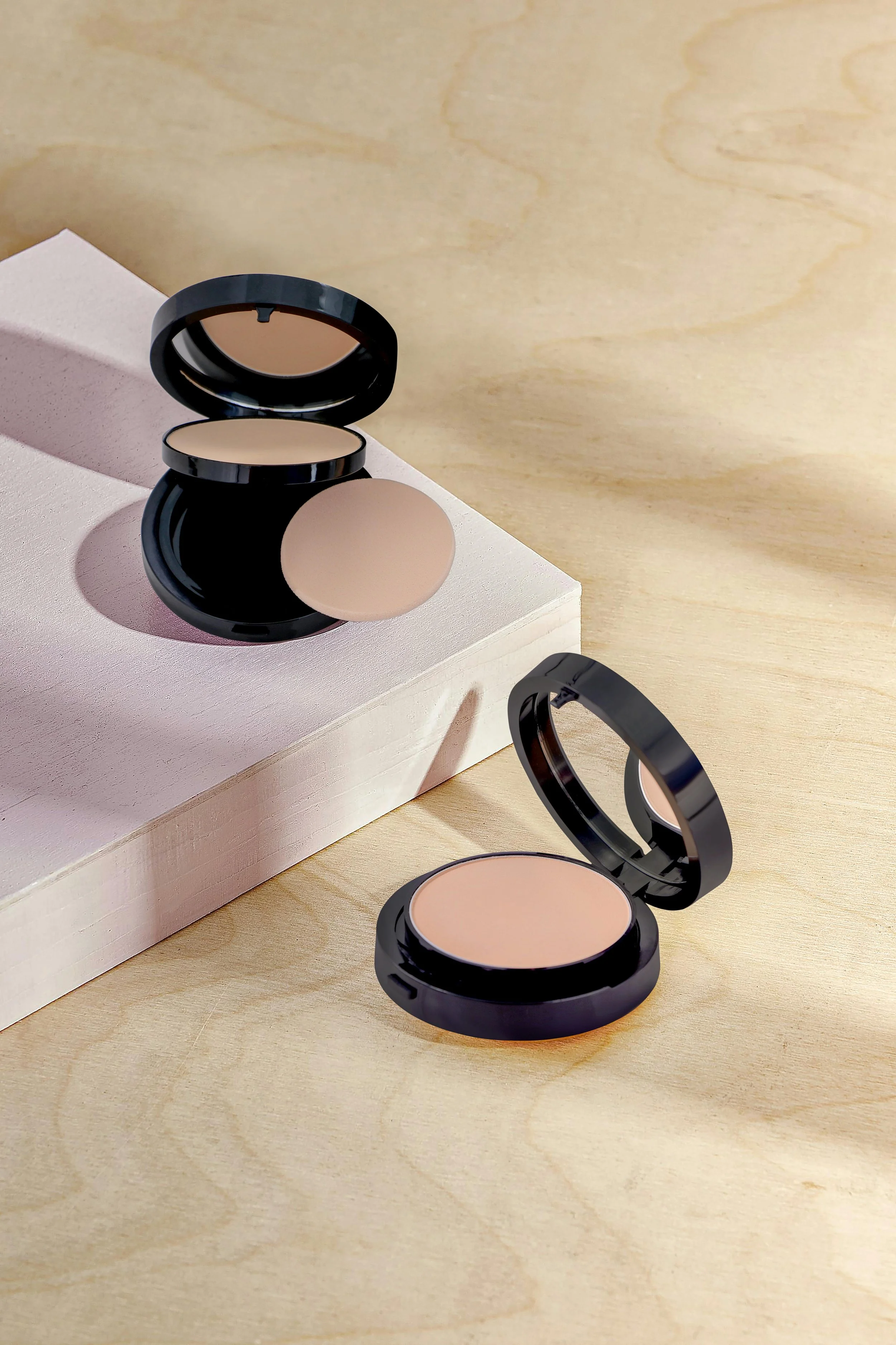 Open makeup compacts with beige powder inside, placed on a light wooden surface and a white rectangular block.