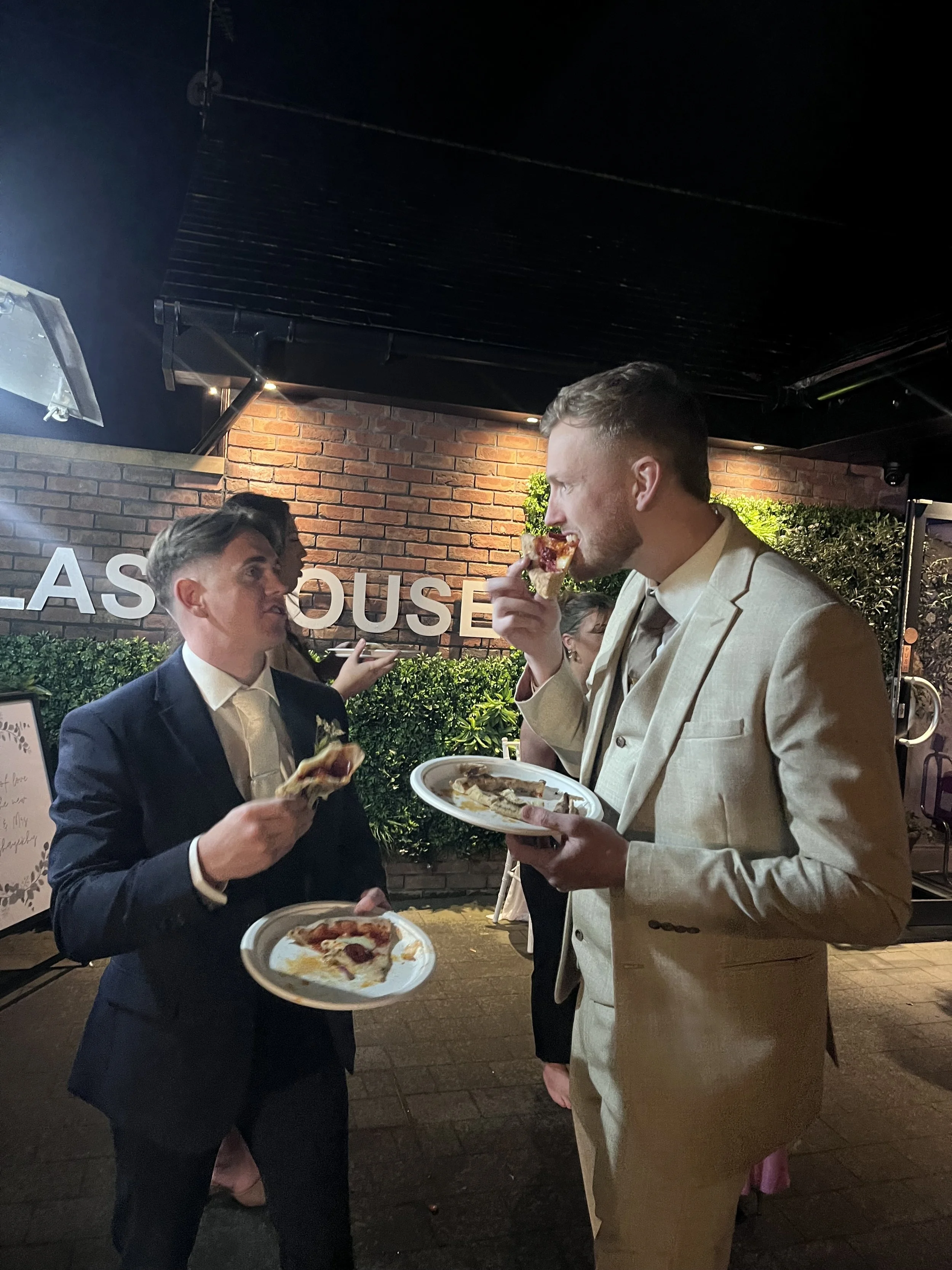 Two men in suits eating pizza at an outdoor event at night.