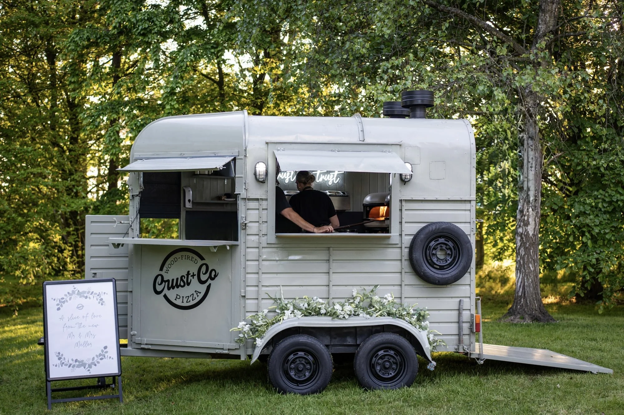 A food truck named 'Crust + Co' selling wood-fired pizza, set in a park with green grass and tall trees in the background, and a sign in front with a message to M & M Mellers.