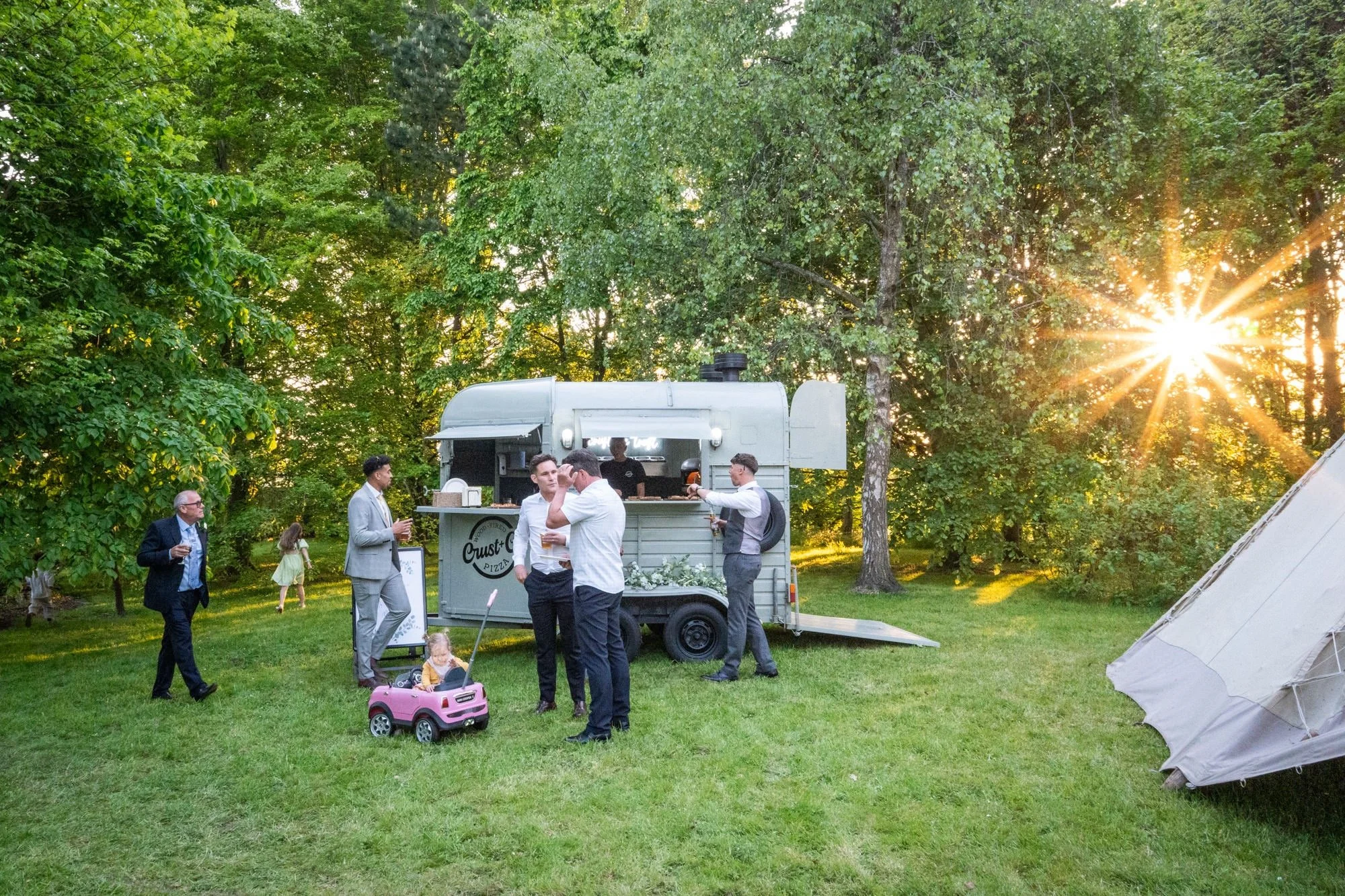 People gathered outdoors near a food truck, with a child in a pink toy car, trees, and sunlight in the background.