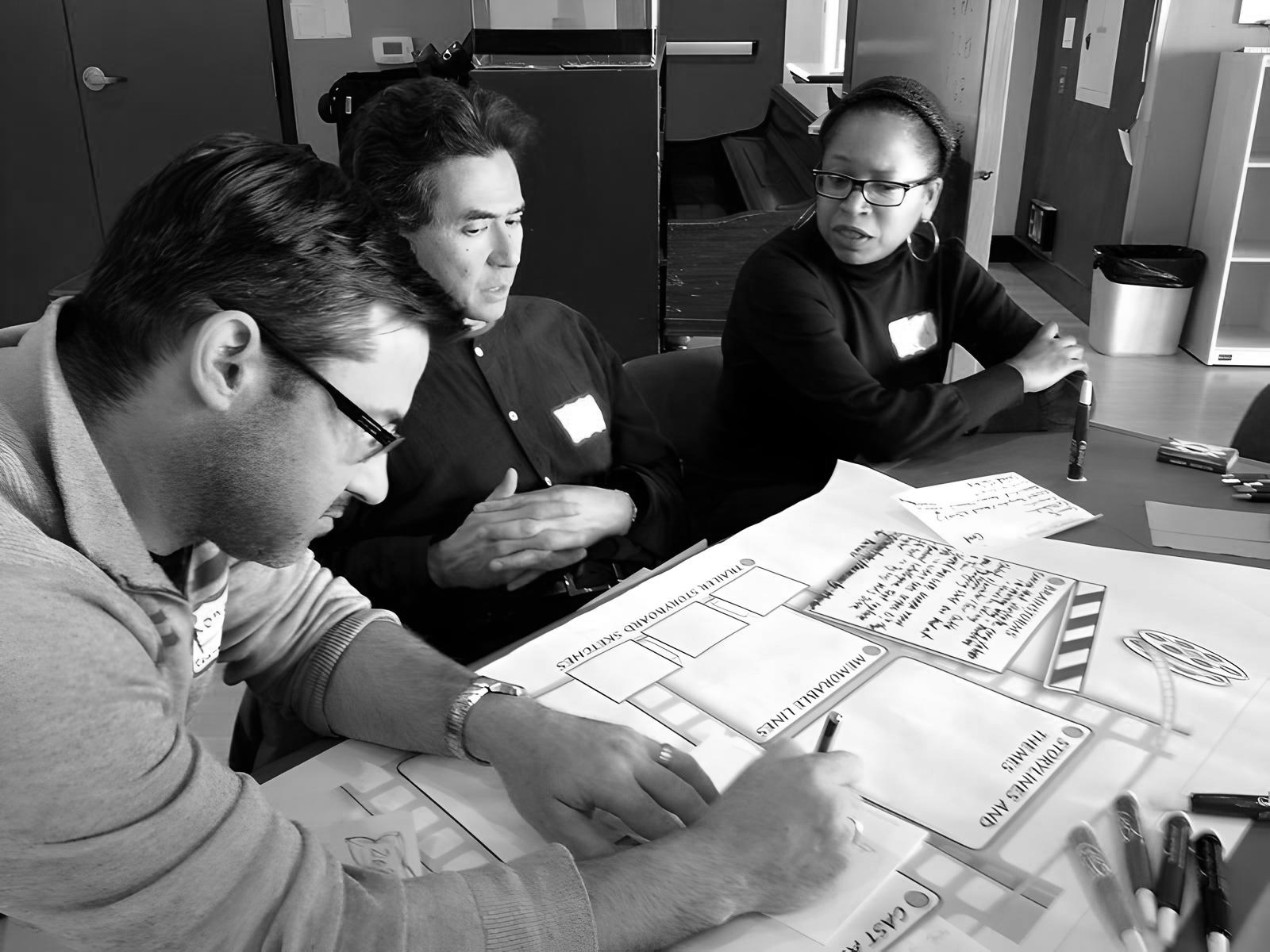Three people collaborating at a table with charts and notebooks, working on a creative project or plan.