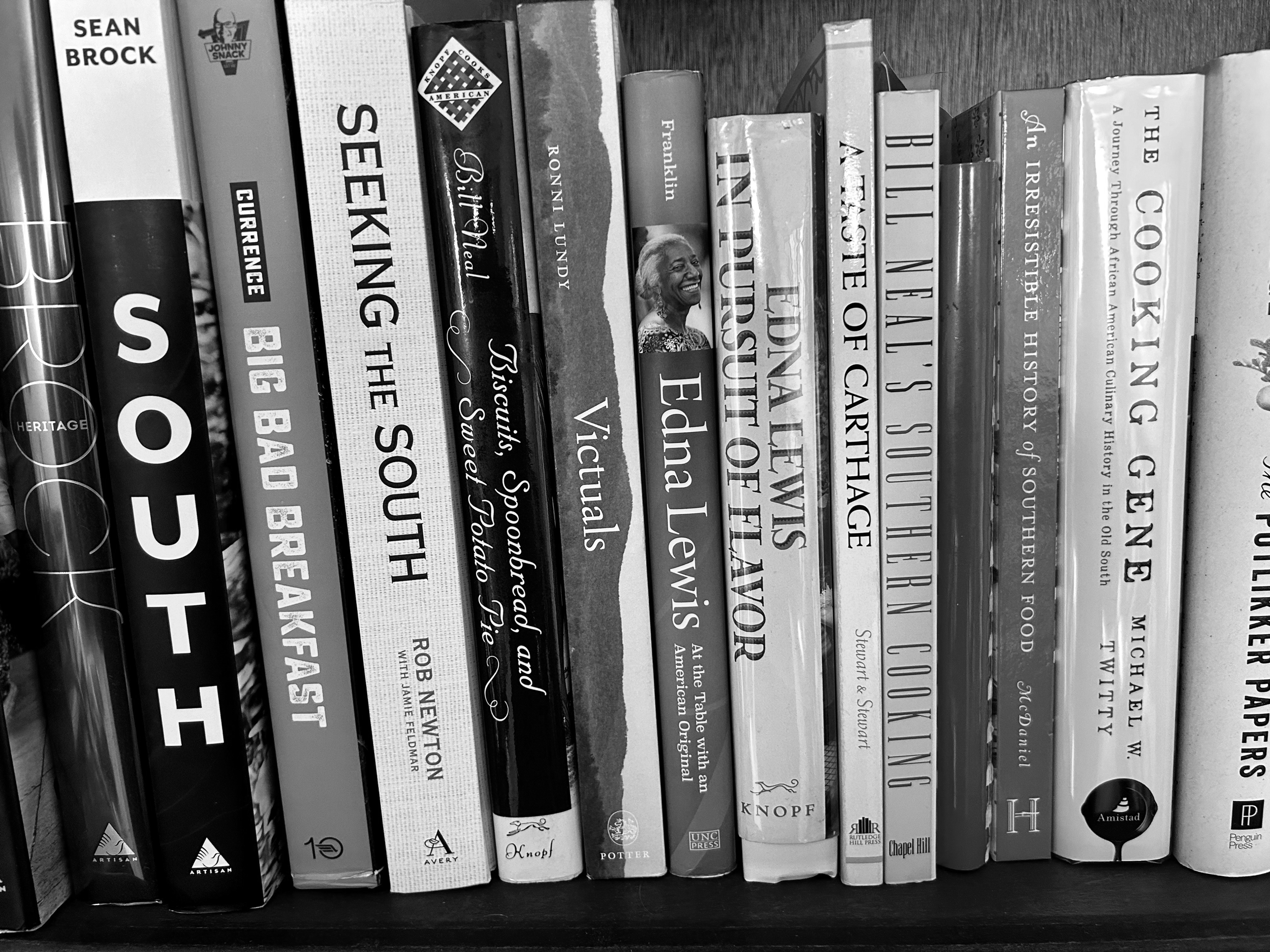 Books on a shelf about Southern cooking and culture, including titles like "South," "Big Bad Breakfast," "Victuals," "Edna Lewis: In Pursuit of Flavor," and "The Cooking Gene."