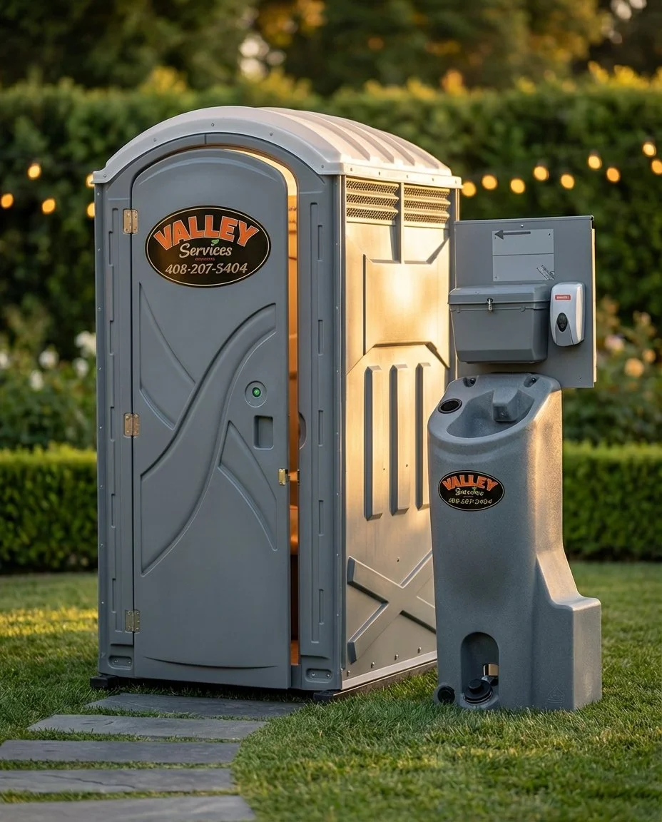 A portable restroom and a hand sanitizing station situated outdoors on grass, with a hedge and string lights in the background.
