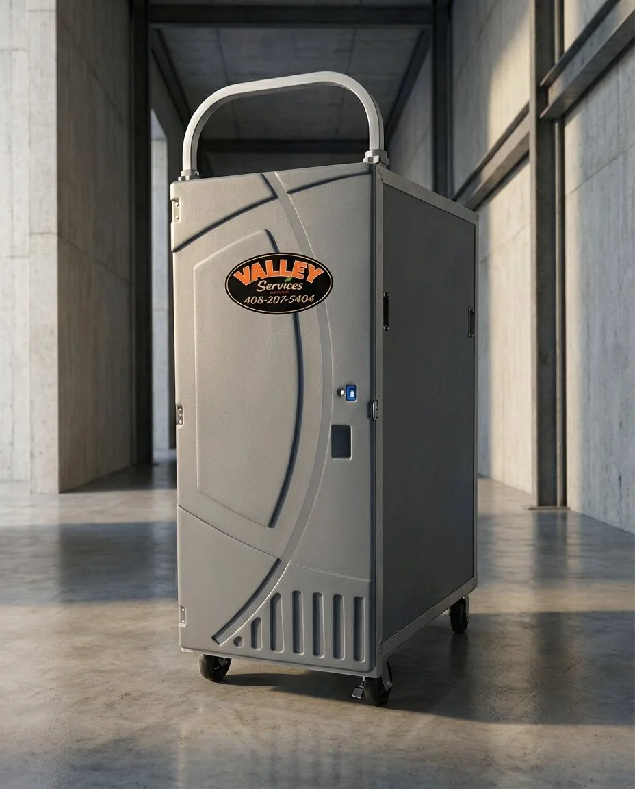A gray portable air conditioning unit with black and orange Valley Services logo on the front, positioned in a modern indoor space with concrete floors and walls.