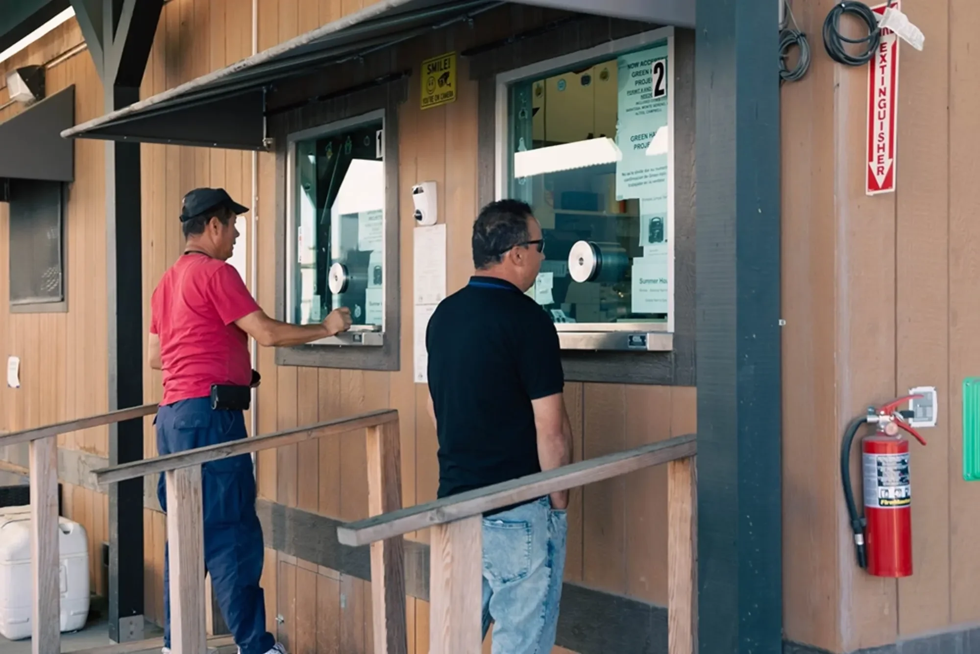 Two people at ticket booth windows, wooden building exterior with a fire extinguisher on the wall.