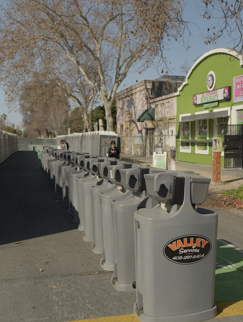 A row of gray electric scooter charging stations on a sidewalk, with a few people near them, and a colorful restaurant building with a green facade in the background, leafless trees, and a clear blue sky.