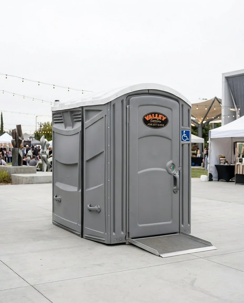 A portable wheelchair accessible toilet at an outdoor event with tents, string lights, and people in the background.