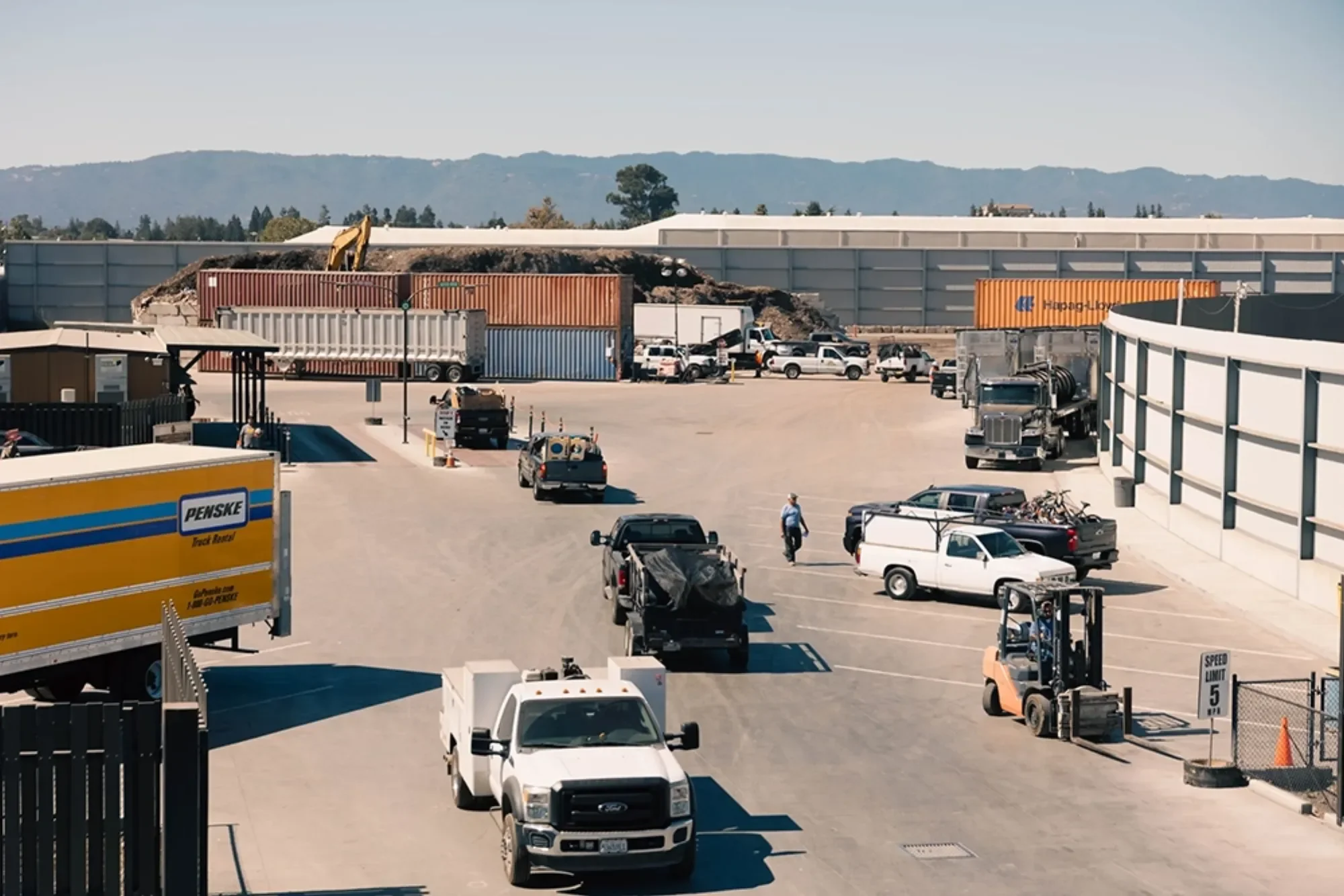 A parking lot with various trucks, cars, and a forklift, with a construction area in the background, and a mountainous landscape beyond.