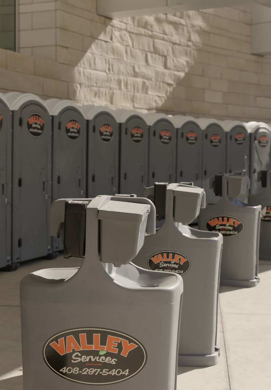 A row of portable hand sanitizing stations with Valley Services logo and contact number, set against a background of gray portable toilets and a stone building.