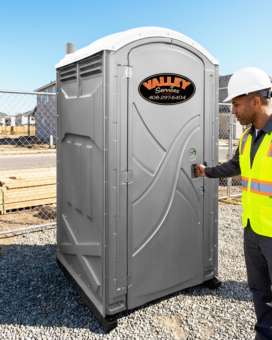 Construction worker in a hard hat and safety vest operating a portable toilet at a construction site with fencing and buildings in the background.