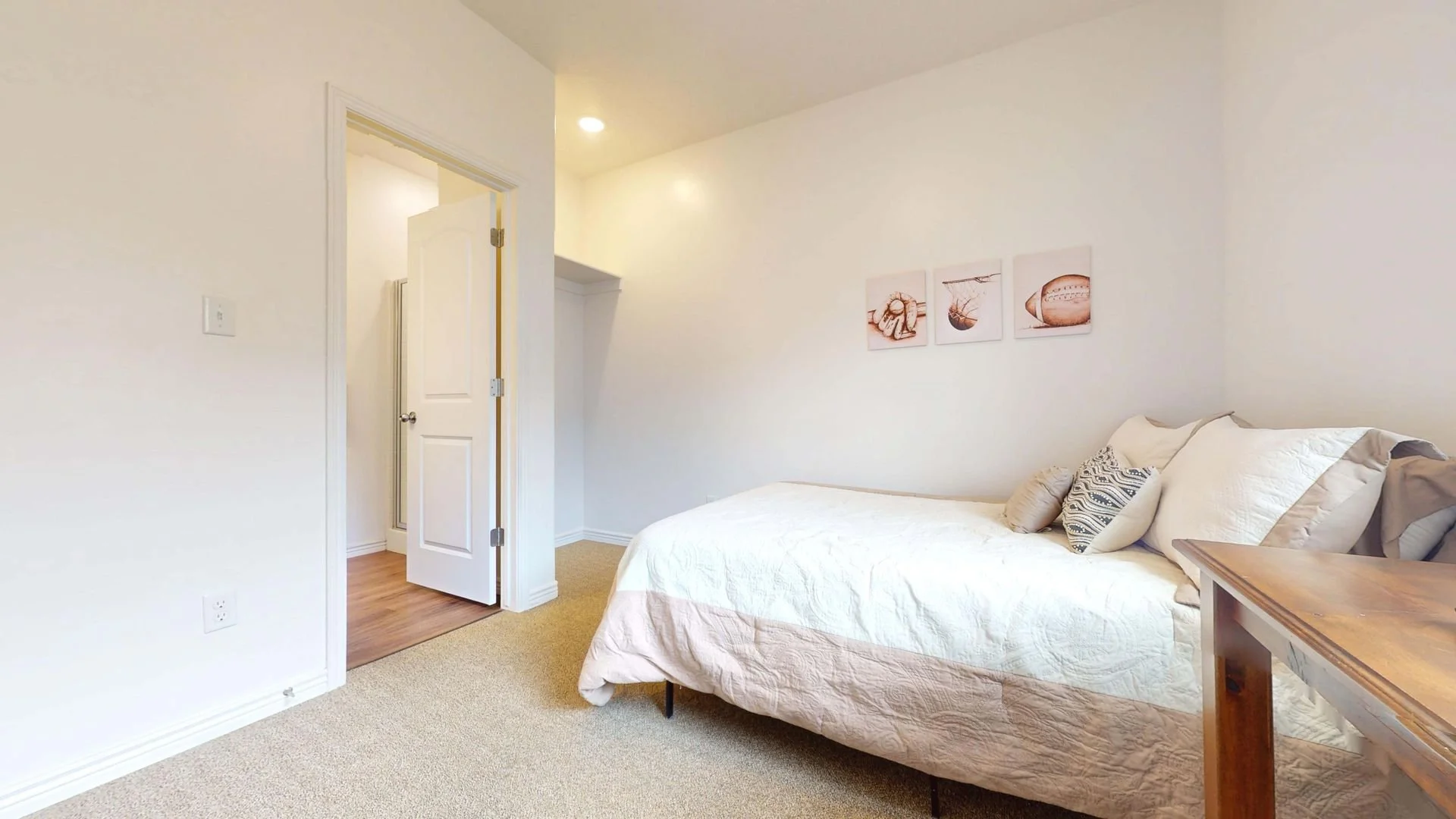 A bedroom with a bed, pillows, and a wooden side table, with a doorway leading to a bathroom, and a wall decoration of three framed pictures of sports equipment