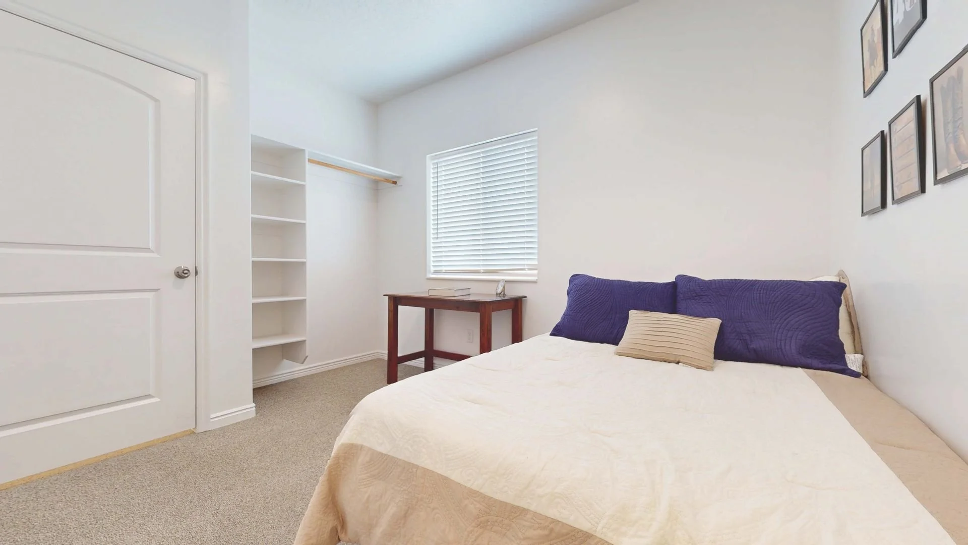 A simple bedroom with a bed, purple pillows, a beige bedspread, a window with blinds, a small wooden desk with books, an empty white bookshelf, a white door, and framed pictures on the wall.