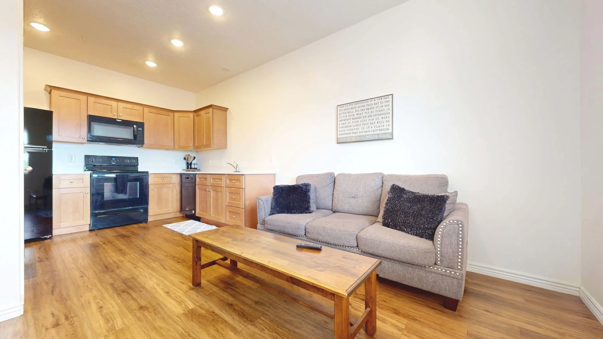 Living room with a beige sofa, black pillows, wooden coffee table, and an open kitchen with wooden cabinets, black appliances, and a tile floor.