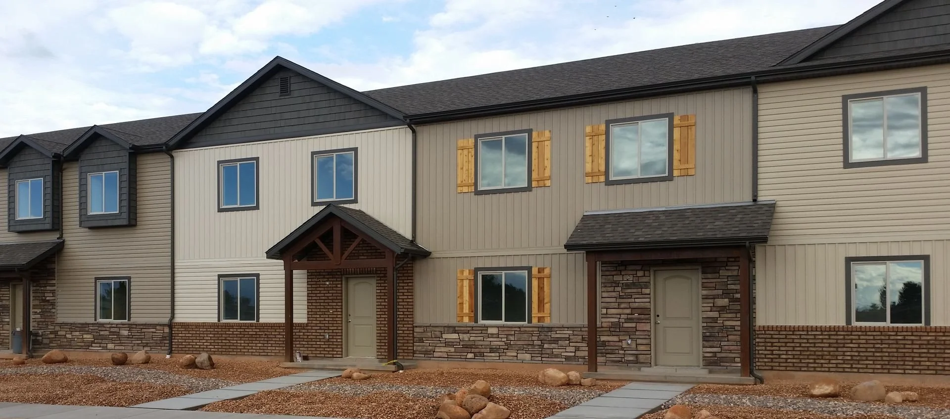 Newly constructed row of townhouses with beige and dark gray siding, stone accents, small covered entryways, and windows with protective wooden shutters, under a partly cloudy sky.